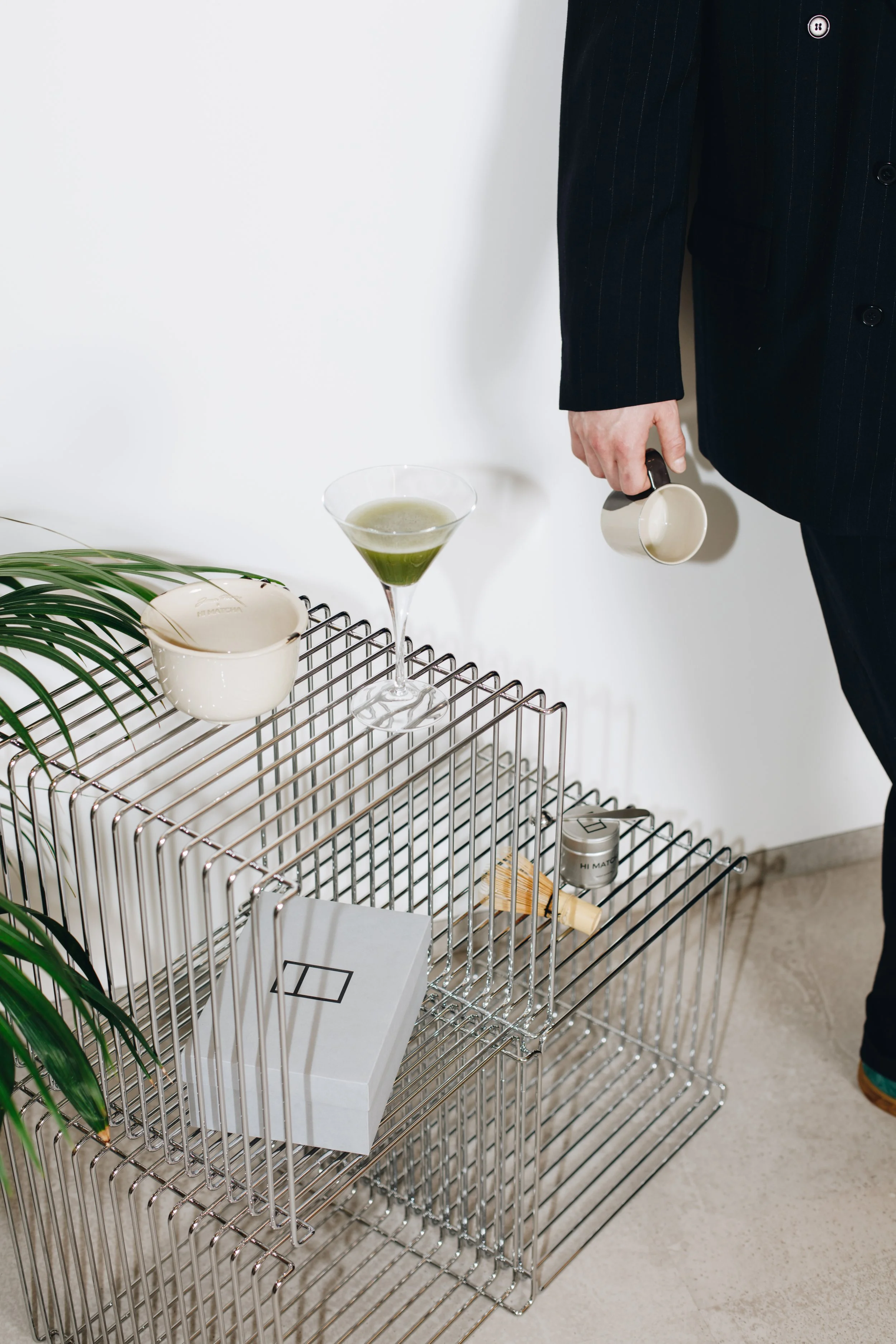 Person in a dark suit holding a white coffee mug, standing next to a modern wire shelving unit with various items, including a box, a small container, a wooden stick, a potted plant, a white mug, and a cocktail glass with a green drink, against a white wall.