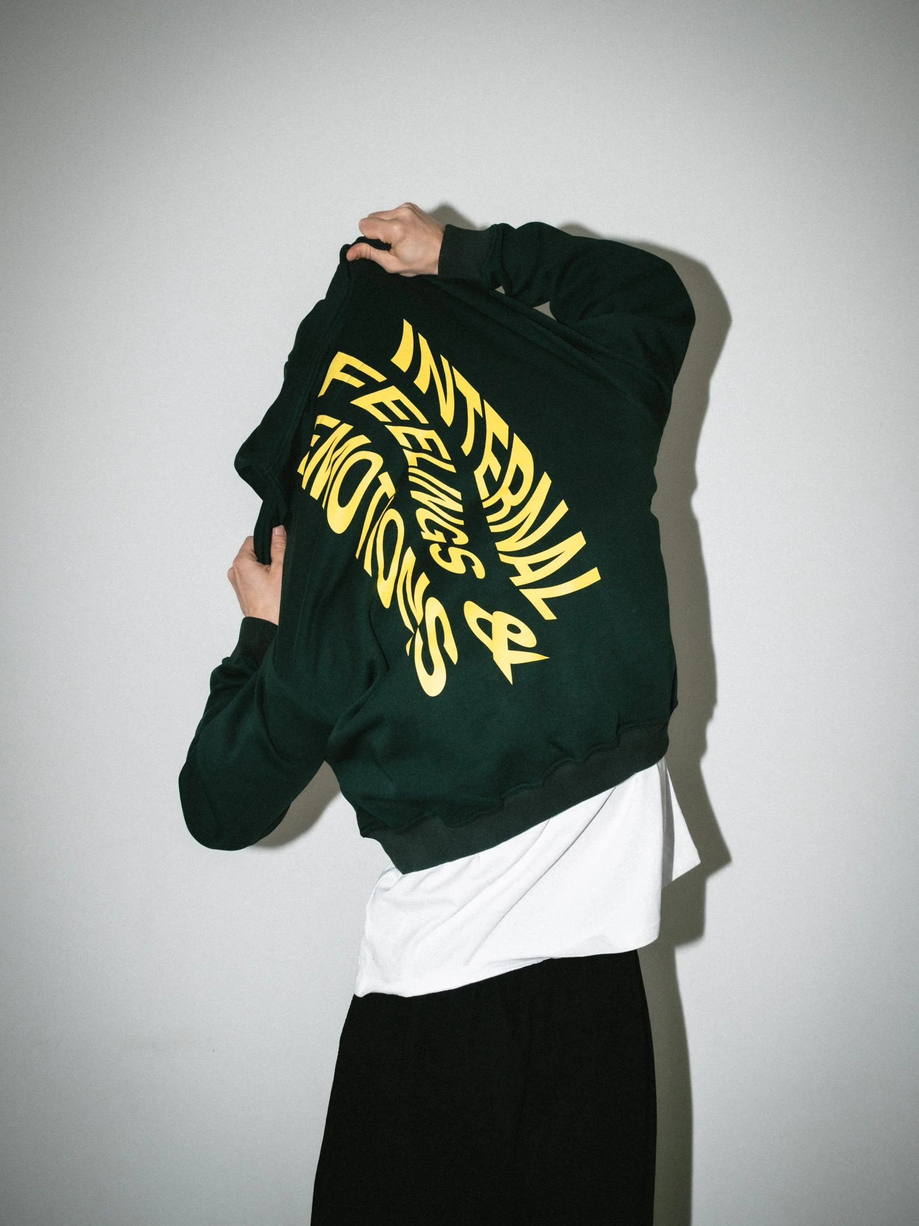 Person wearing a black hoodie with yellow text, lifting the hood while standing against a plain white wall.