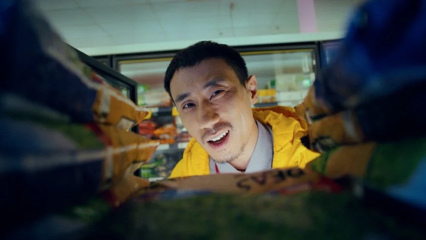 A man looking down into a grocery store aisle, surrounded by stacked snack bags, wearing a yellow jacket.