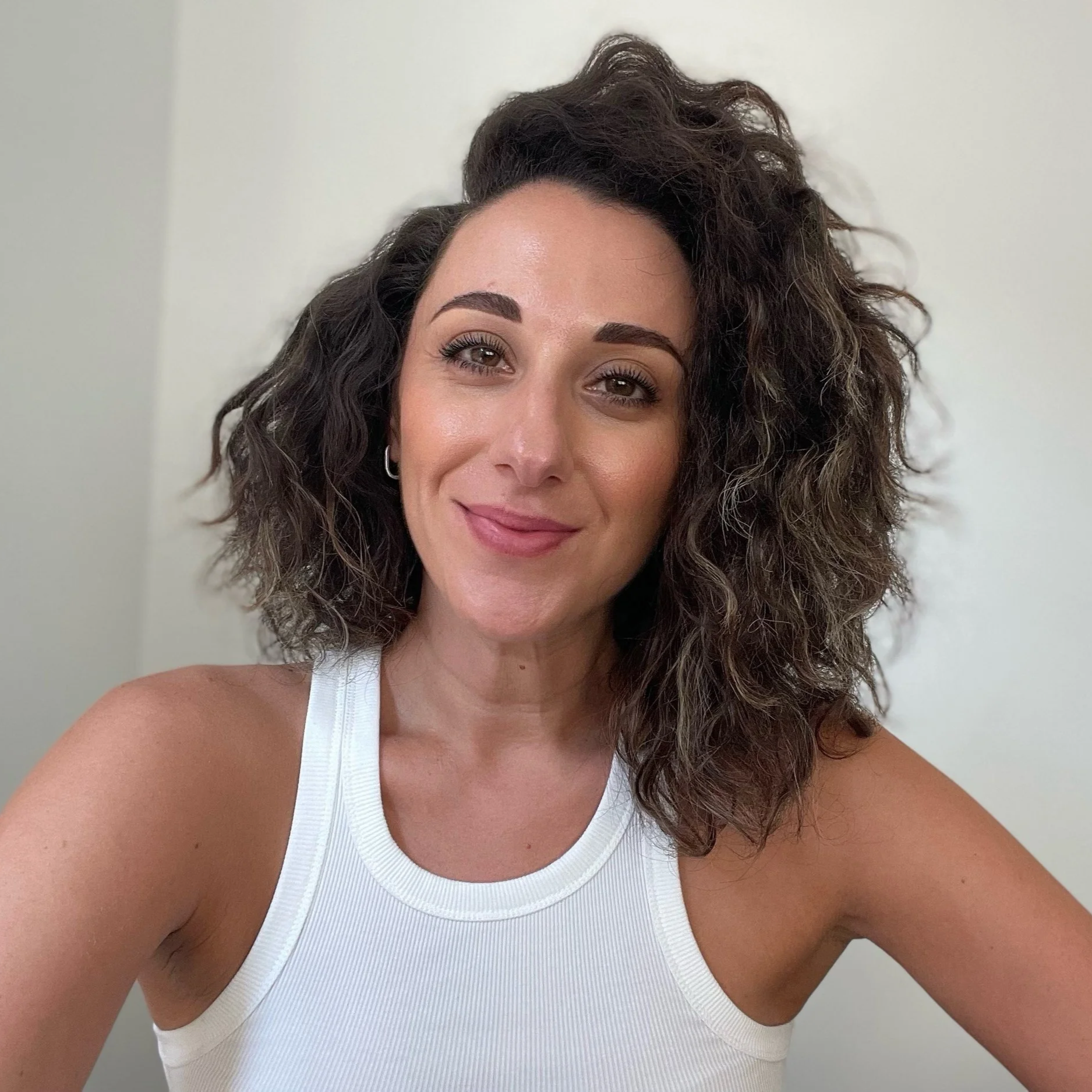 A woman with curly dark hair, wearing a white tank top, smiling at the camera.