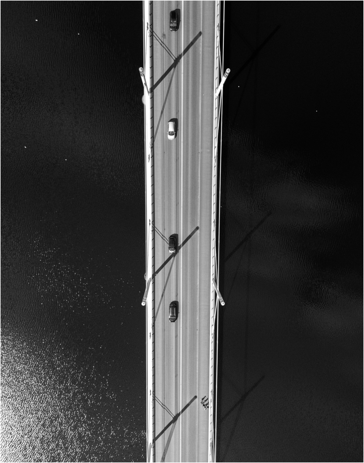 An aerial black and white photo of a bridge spanning over water, with cars driving across it. The bridge has four large light poles and shadows cast on the road.