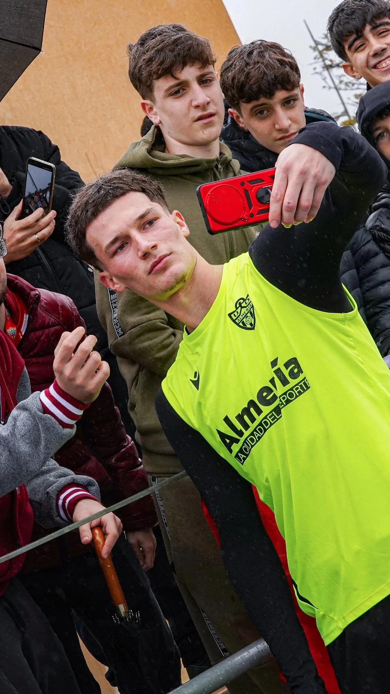Un grupo de jóvenes toma una selfie con un joven en primer plano vestiendo una camiseta amarilla con el logo de Almería en un evento al aire libre.