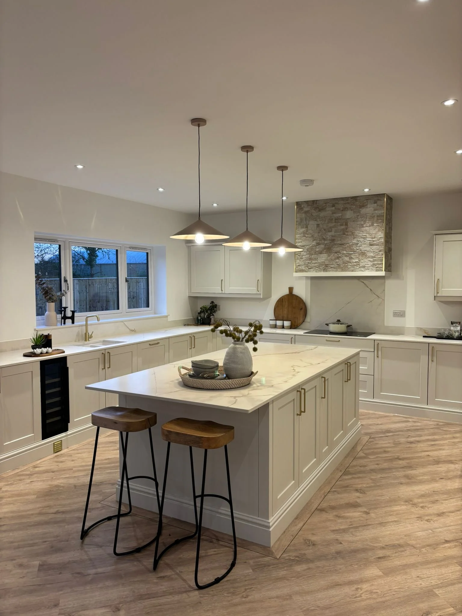 Modern kitchen with white cabinets, marble countertop island, wooden bar stools, pendant lights, view of the window, and a stone feature wall.