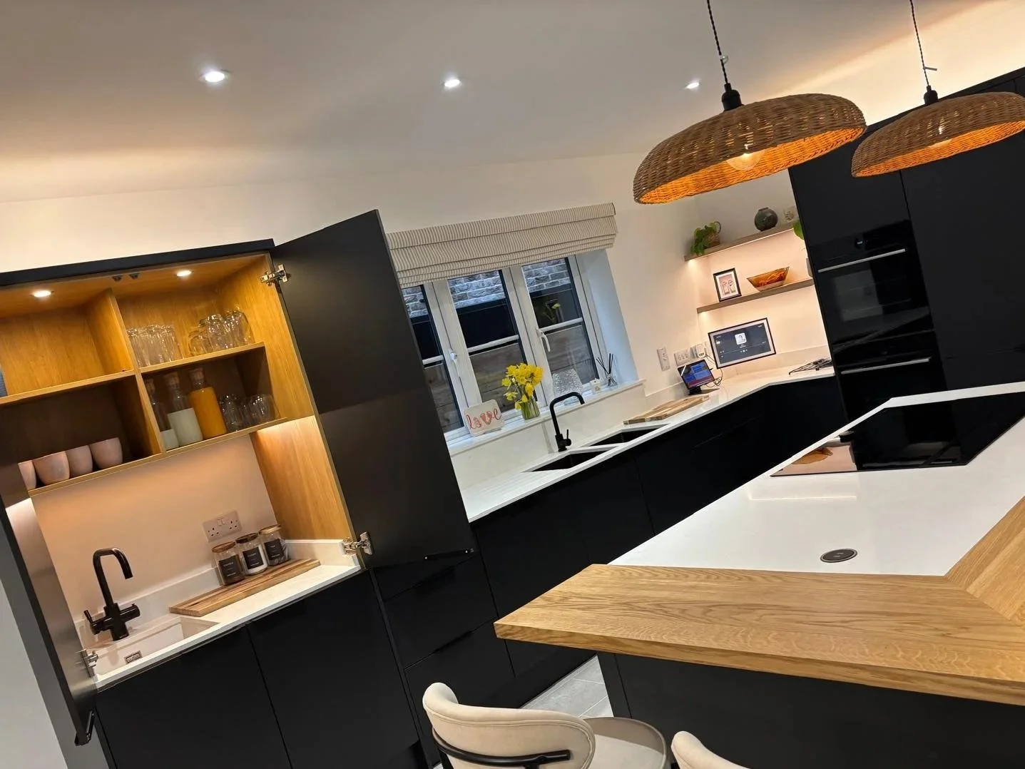 Modern kitchen with black cabinets, white countertops, wooden accents, hanging woven lights, and a window with a vase of yellow flowers.