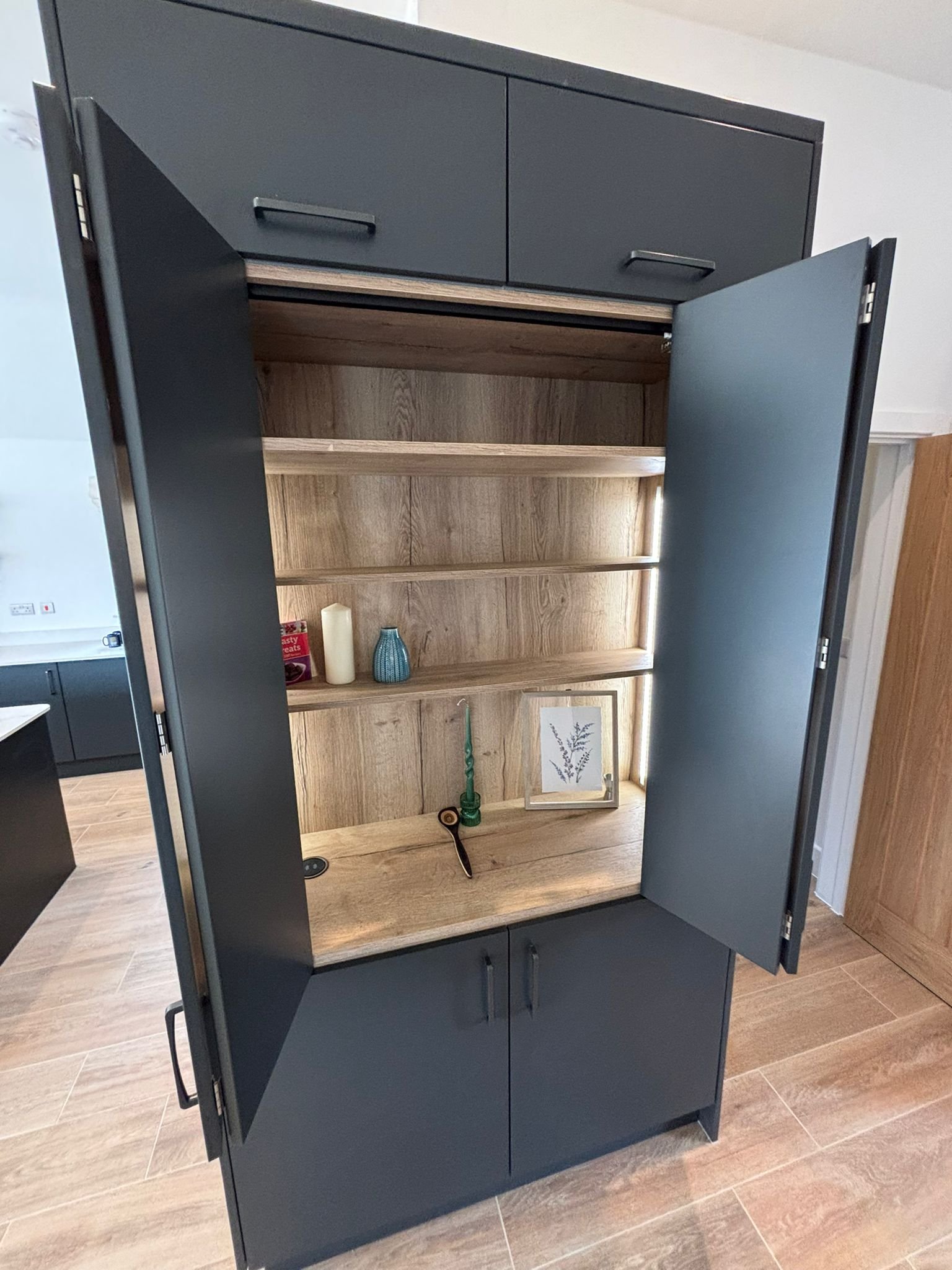 A dark-colored cabinet with open doors revealing a wooden interior with shelves. The cabinet is decorated with a framed botanical print, a green candle, a blue vase, candles, and a snack box on the shelves.