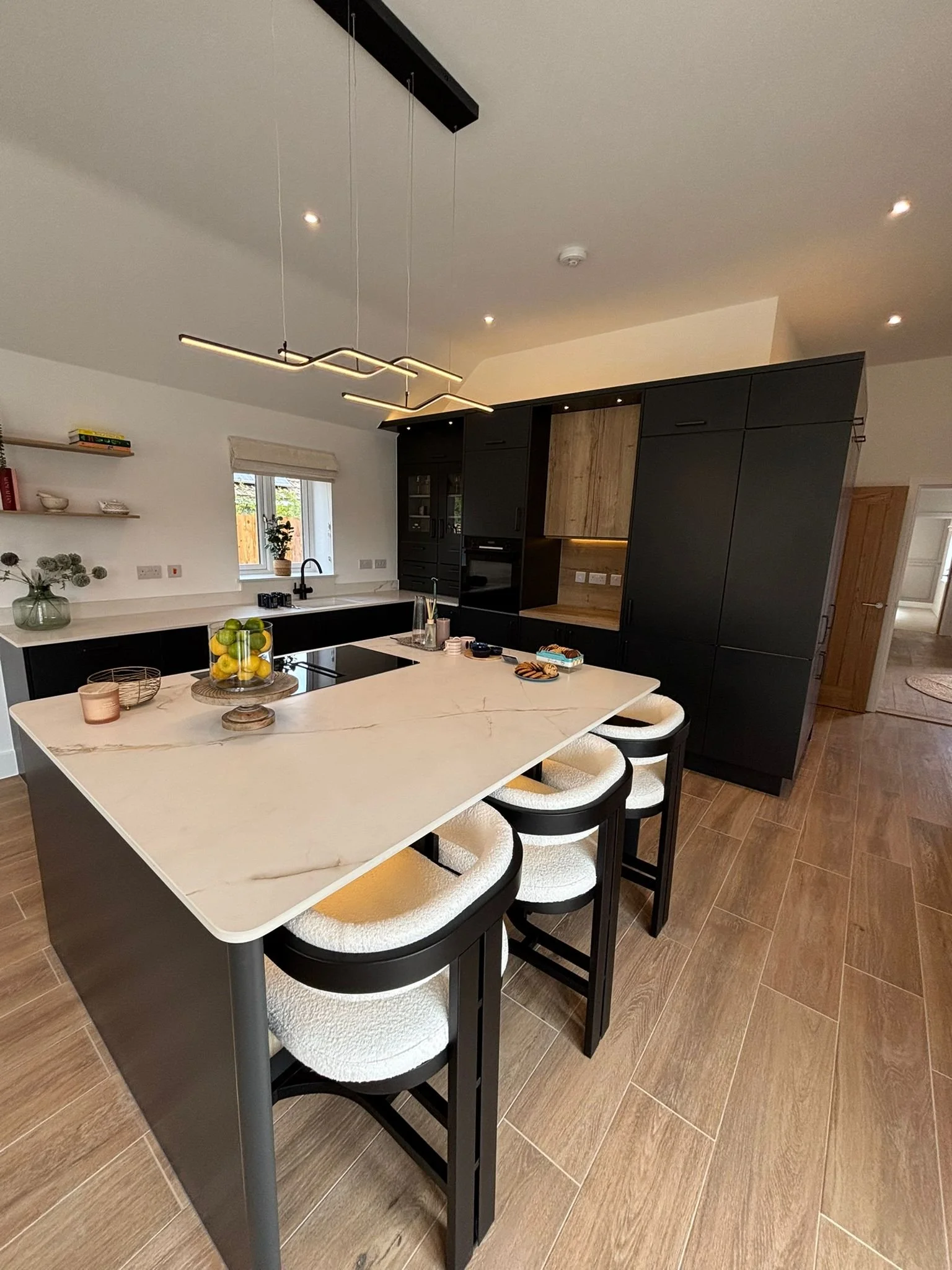 Modern kitchen with white countertop, black cabinetry, and a wooden accent wall, featuring a central island with barstools, a window with a plant, and contemporary lighting.