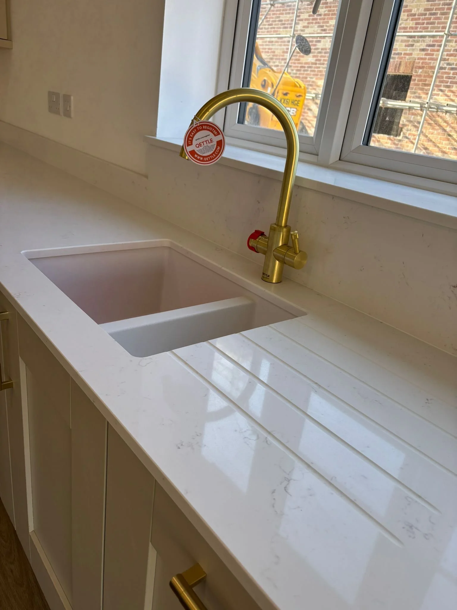 A kitchen countertop with a built-in sink and a modern gold Qettle tap, with a window above showing a brick building and a yellow construction machine outside.