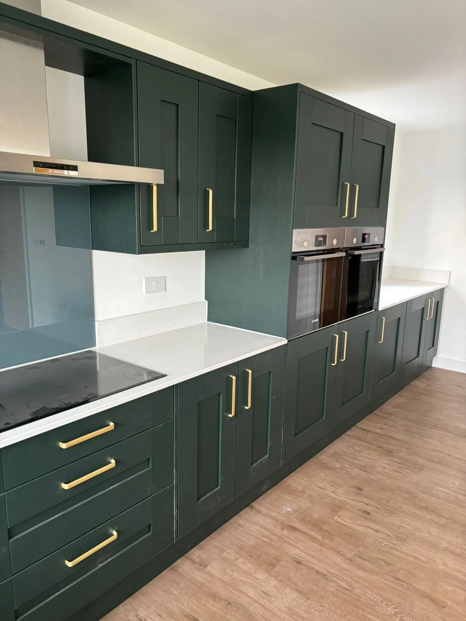 Modern kitchen with dark green cabinets with gold handles, white countertop, wood flooring, and built-in stainless steel oven and microwave.