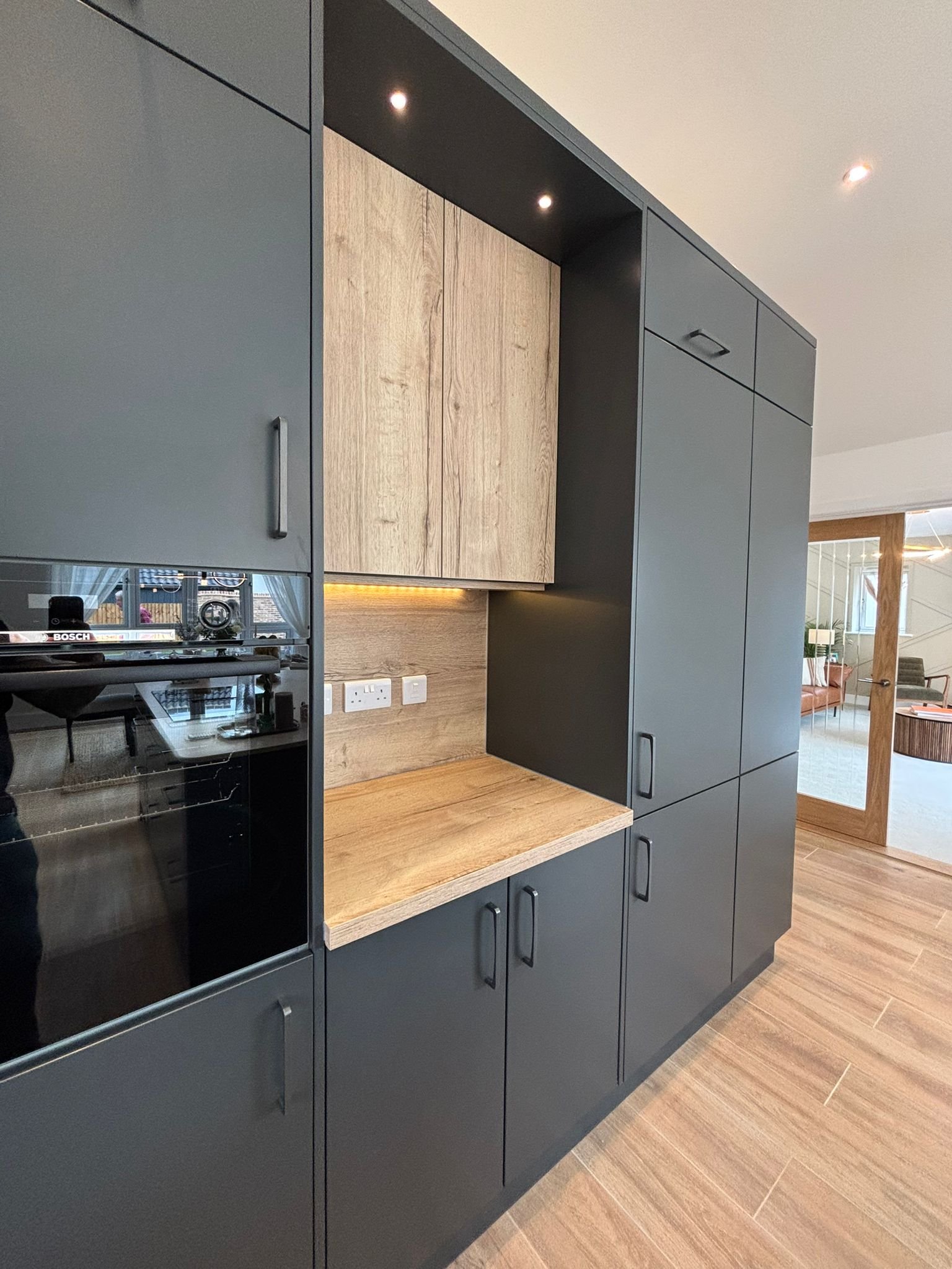 Modern kitchen with dark gray cabinets, wooden countertops, and wooden flooring. Wall outlets and electrical switches are visible below the cabinets. In the background, a living area with a glass door, wooden frame, and some furniture is seen.