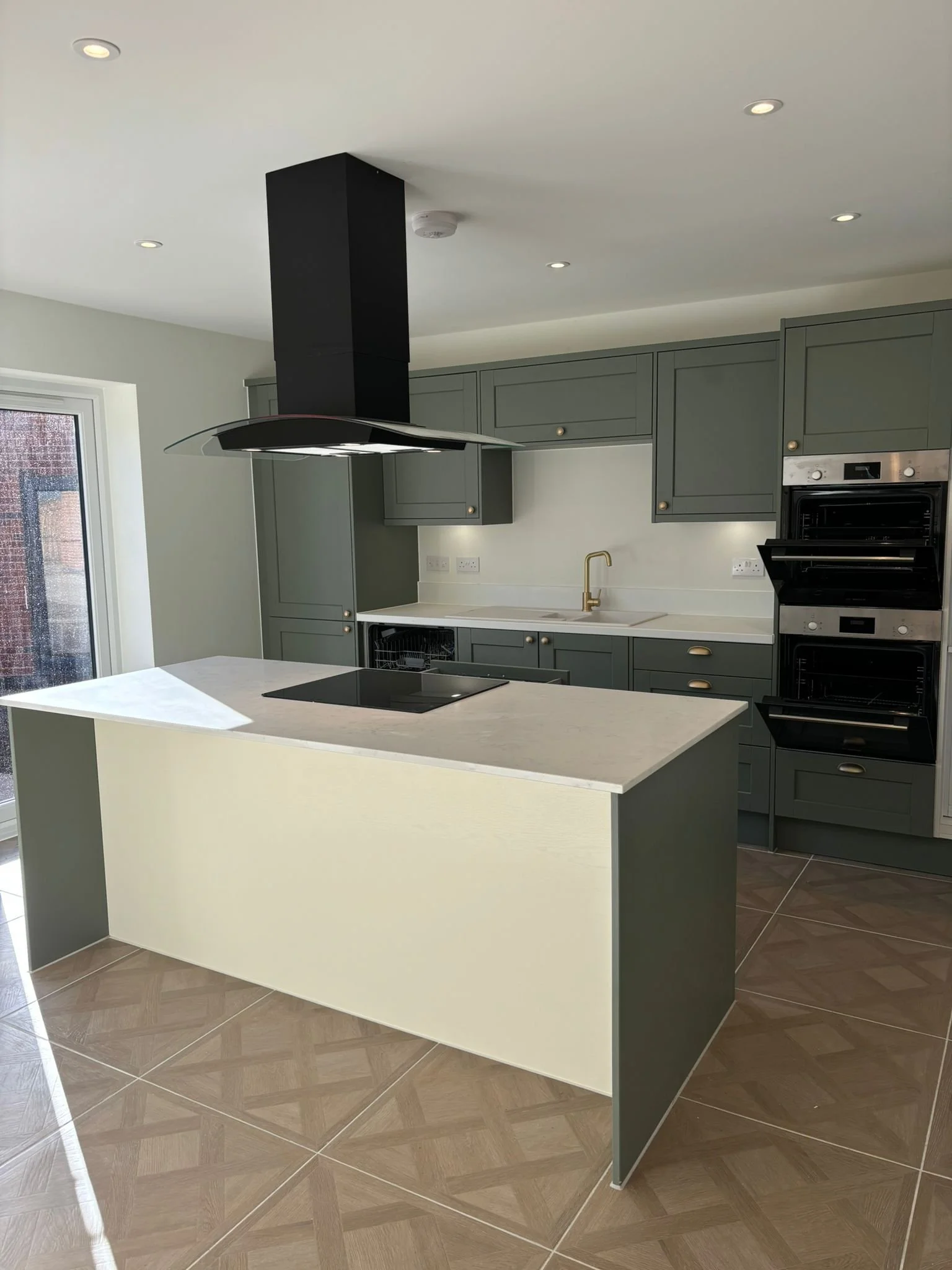 Modern kitchen with grey cabinets, white countertops, a black and gold faucet, and a central island with a stove. There is a built-in oven and microwave, a range hood, and tile flooring. A sliding glass door is visible on the left.