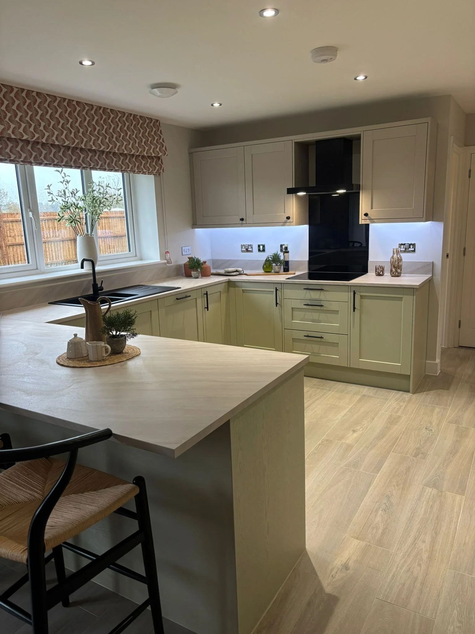 Modern kitchen with beige cabinets, black induction stove, white countertop, window with patterned valance, and decorative vases and plants.