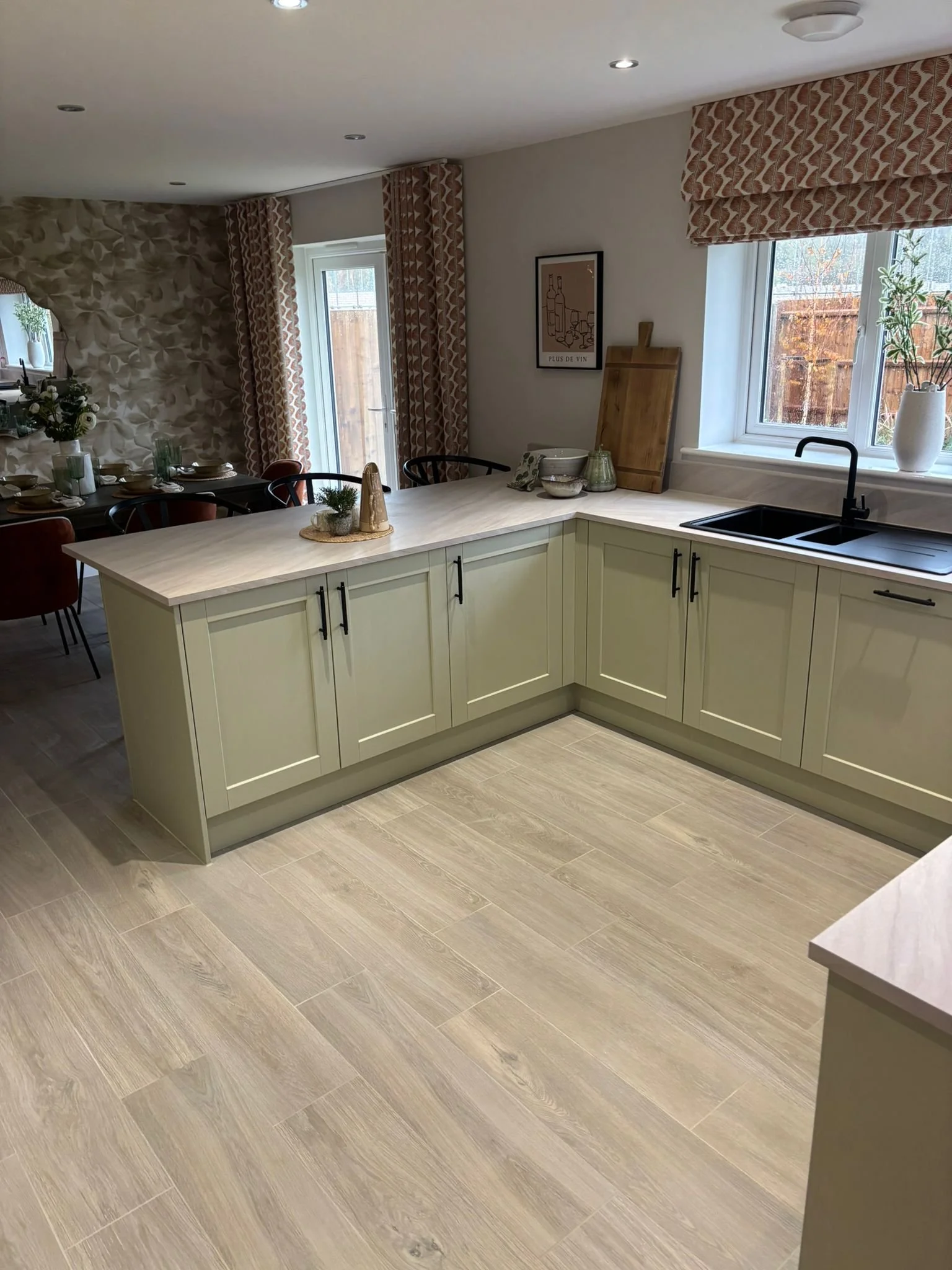 View of a modern kitchen and dining area with cream-colored cabinetry, a black sink, window with patterned Roman shades, dining table with settings, and decorative items on the countertop and wall.