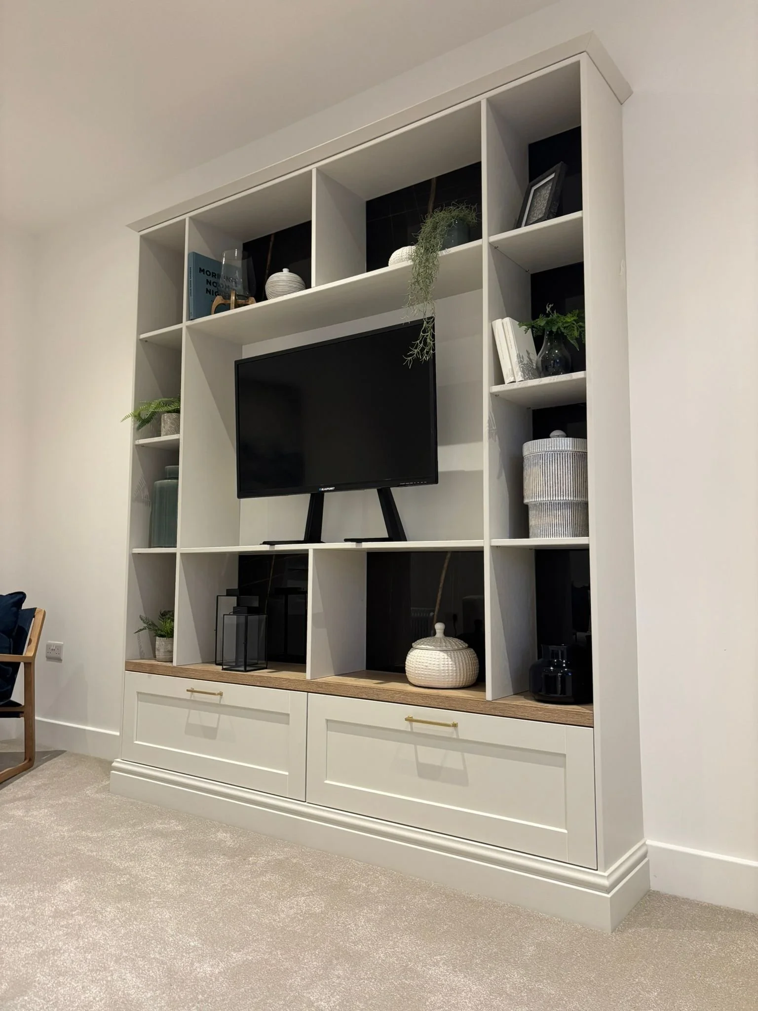 White entertainment unit with decor items, plants, and a mounted flat-screen TV, placed in a living room with beige carpet.