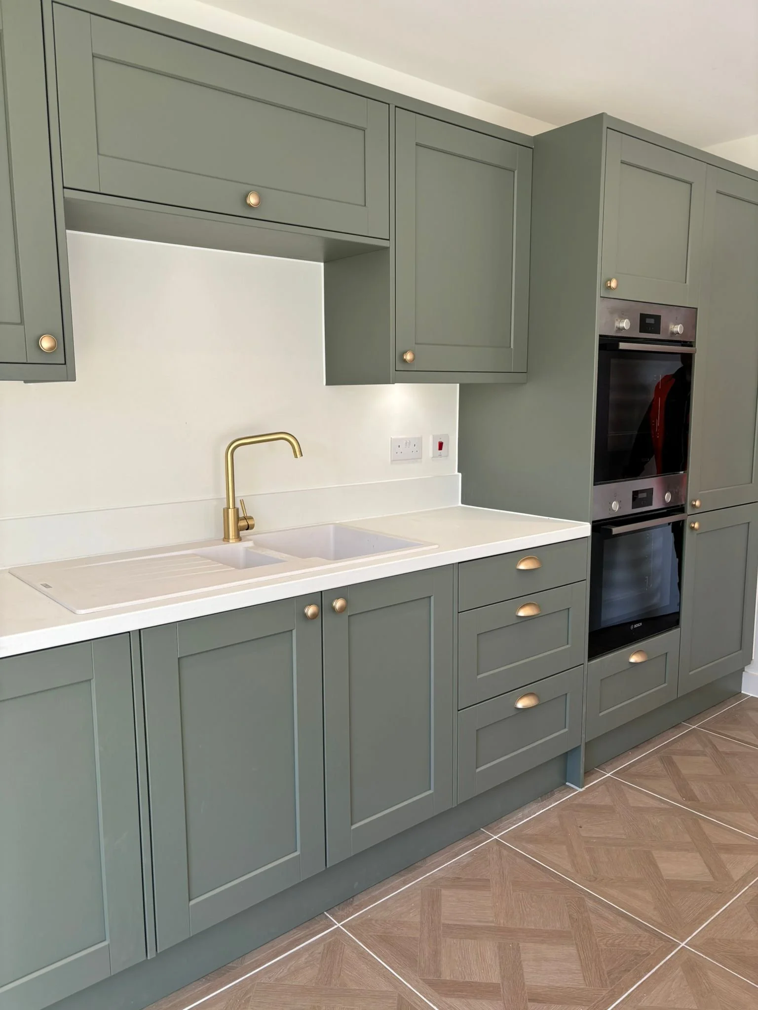 A modern kitchen with green cabinetry, gold knobs, a white countertop, a gold faucet, a built-in oven, and wood-look tile flooring.