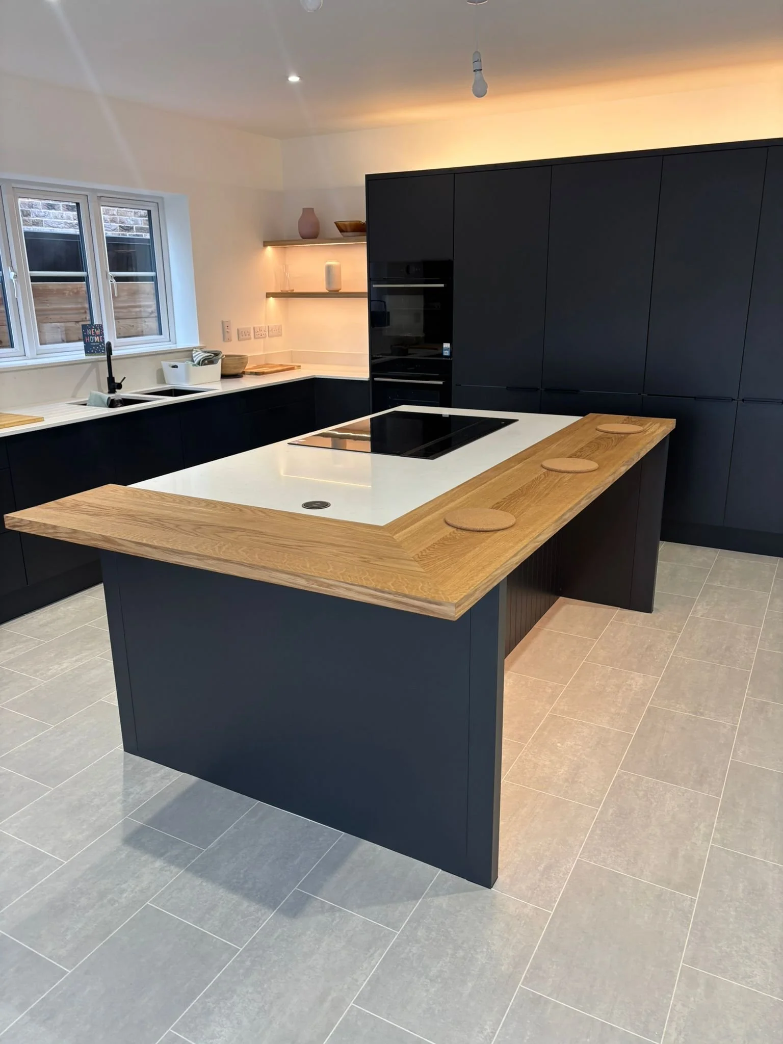 Modern kitchen with black cabinets, a white countertop, and a wooden island with circular wooden coasters. There are shelves with decorative items, a window above the sink, and a tiled floor.