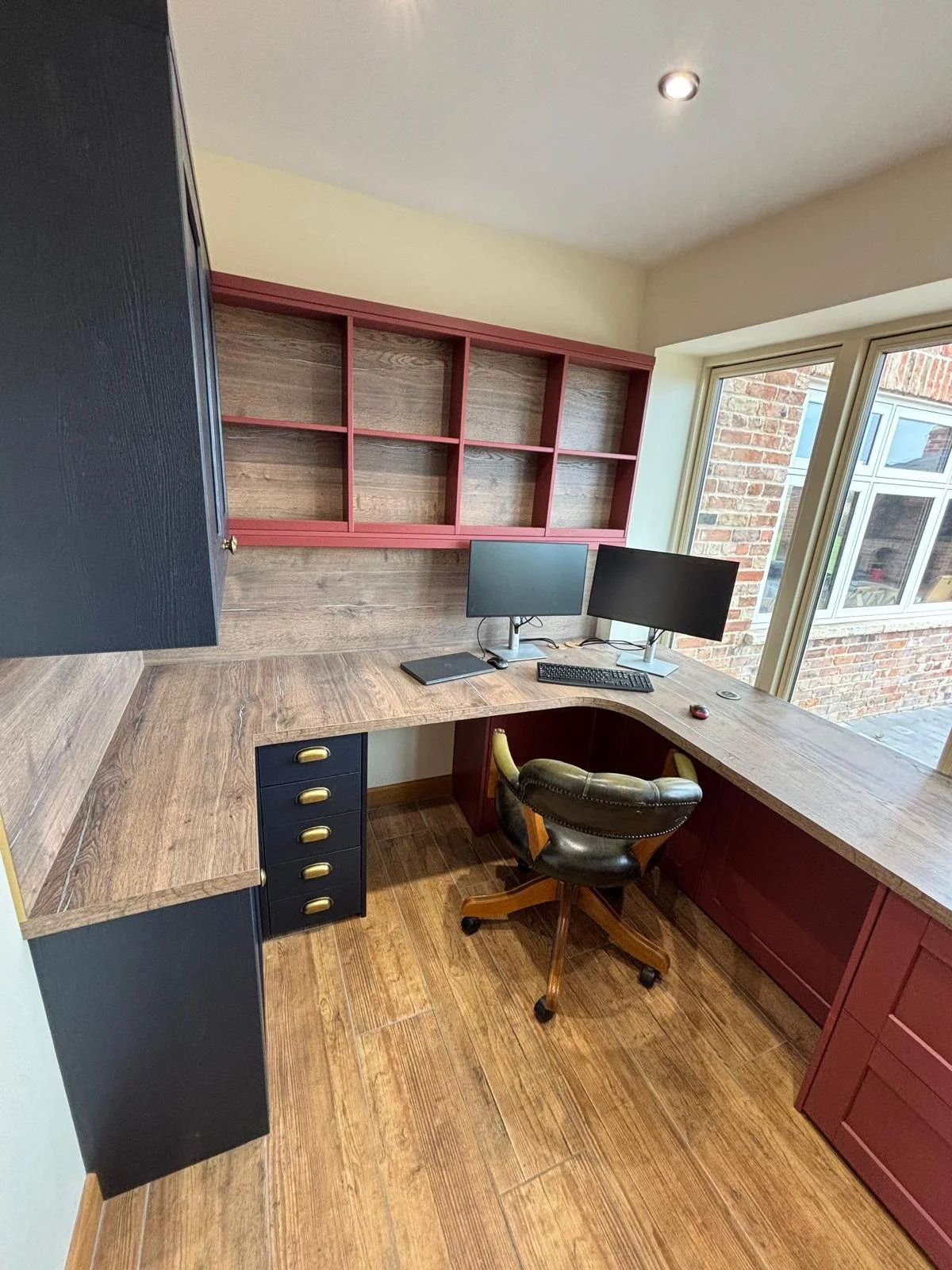 Home office with wooden L-shaped desk, dual monitors, black office chair, brick wall outside window, open red and black shelving