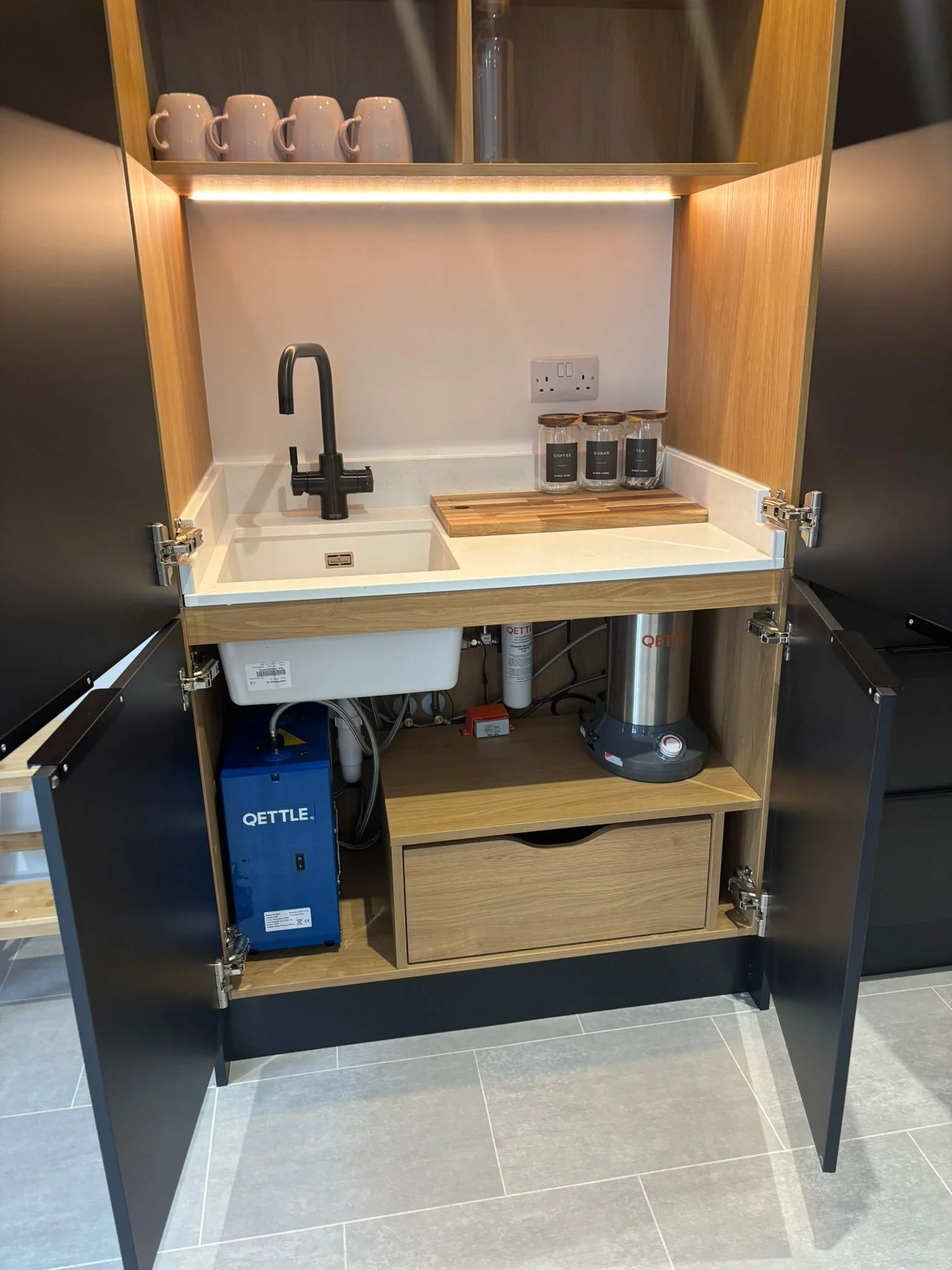 Open kitchen cabinet with a small sink, countertop, and storage space underneath, with a blue water heater, three glass jars labeled coffee, sugar, and tea, and a wooden cutting board on the countertop.