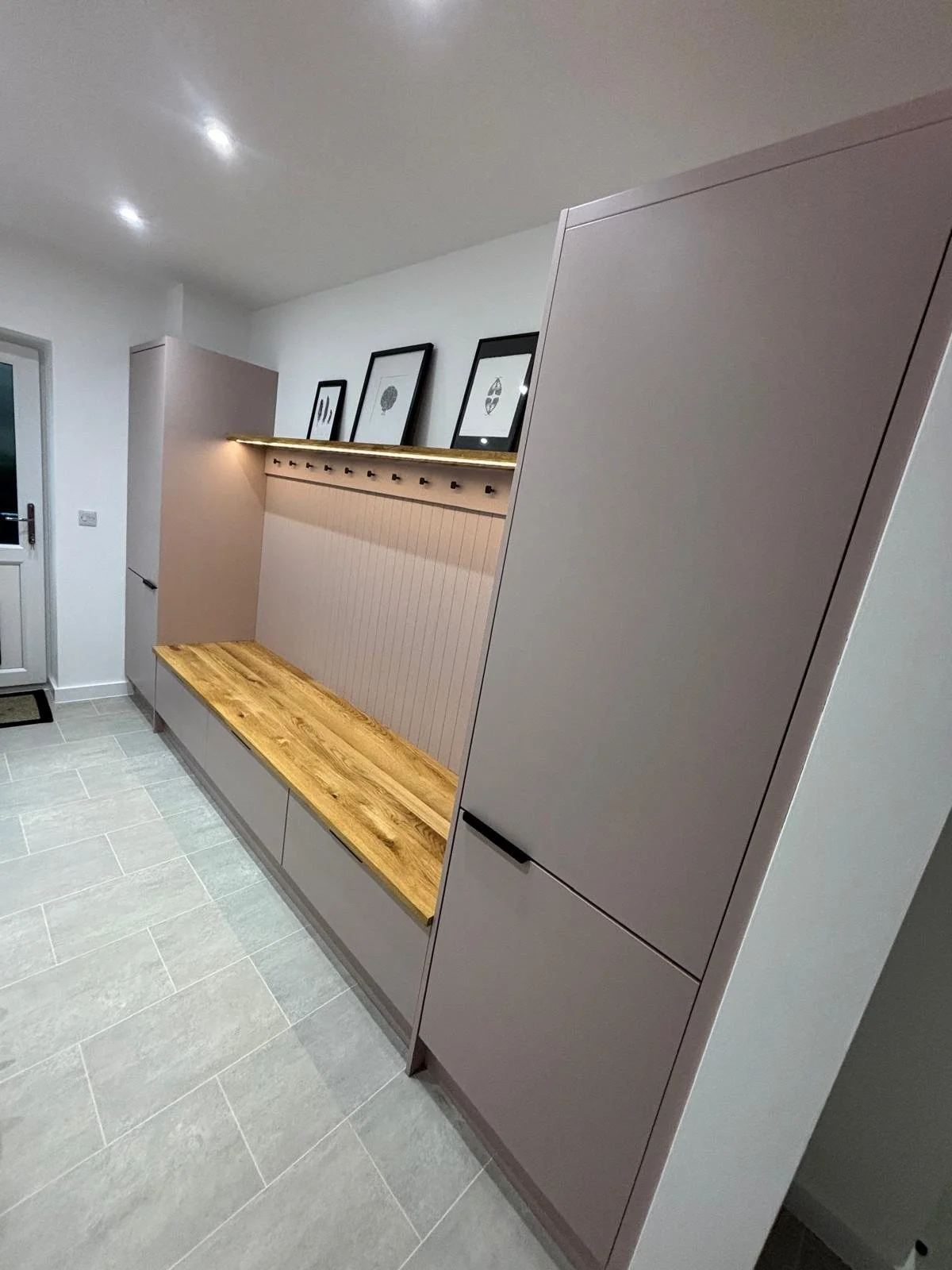 Modern hallway with light gray cabinets, a wooden bench, black-framed artwork, and a white door in the background.