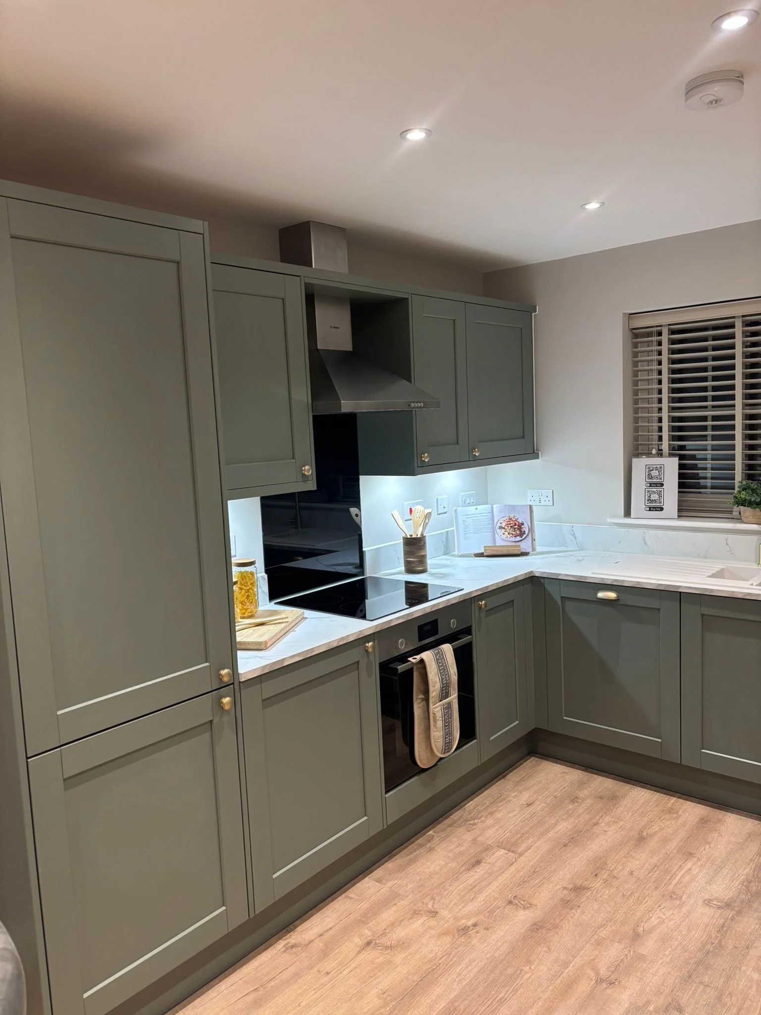 A modern kitchen with green cabinets, a white marble countertop, a built-in oven, a stovetop, and a range hood. There is a window with blinds and various items on the counter, including a jar with utensils and a menu stand.