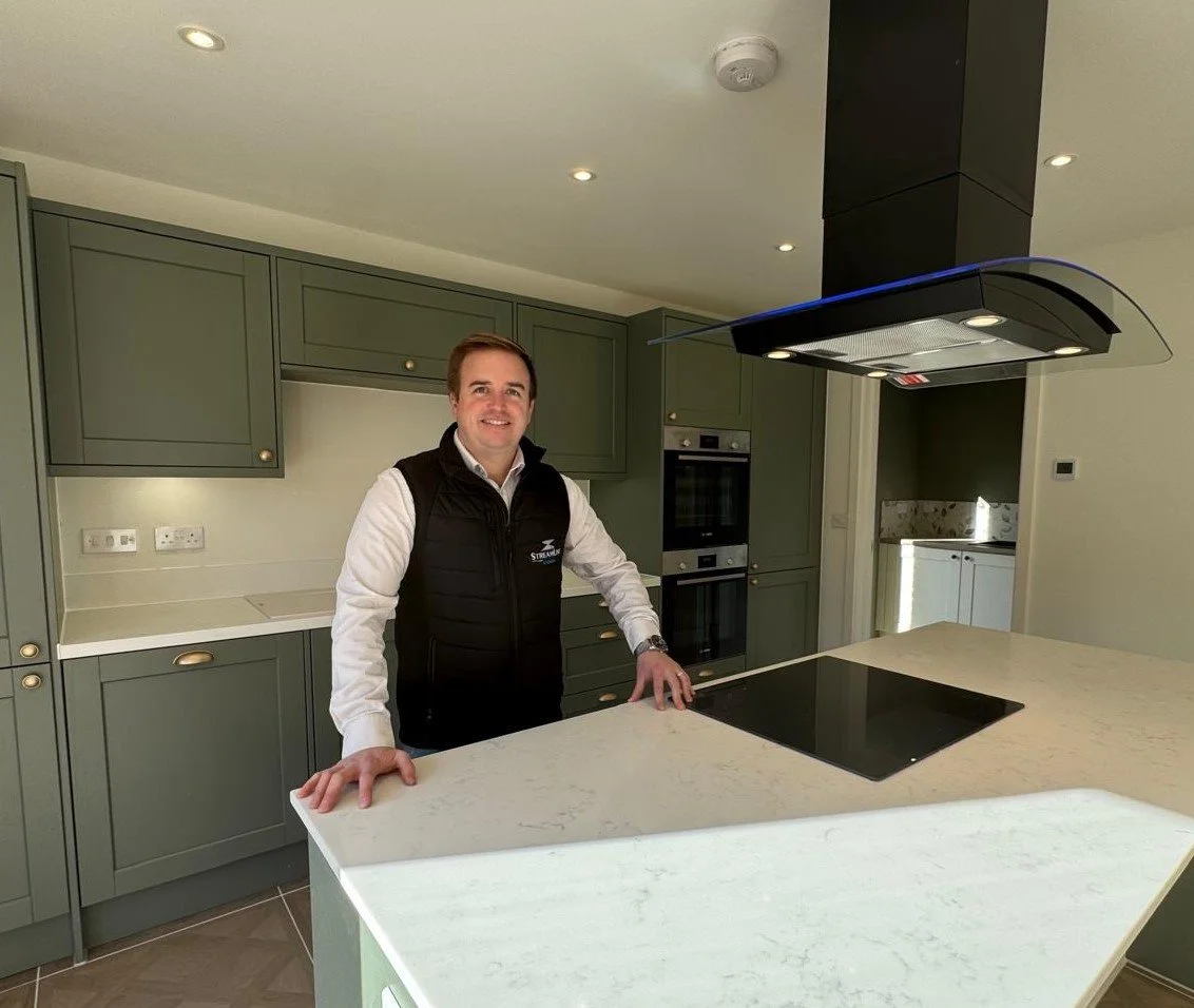 A man standing in a modern kitchen with green cabinets, a white marble countertop, a black stovetop, and built-in oven, smiling at the camera.