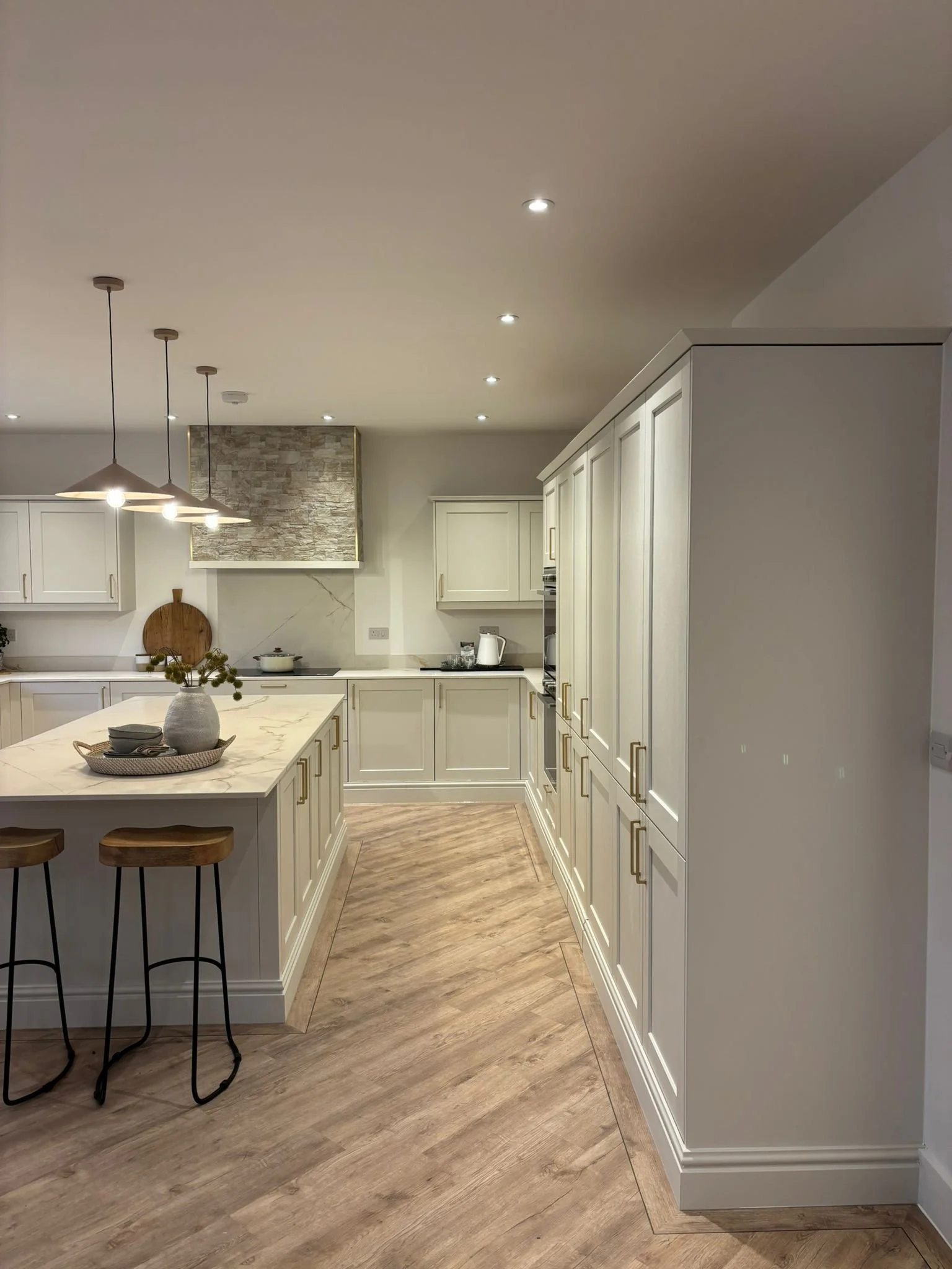 A modern kitchen with white cabinets, a marble island with a vase and bowls, wooden stools, and a wood flooring. There are installed ceiling lights and pendant lights over the island. Part of a stove and a recirculating vent with a stone background a
