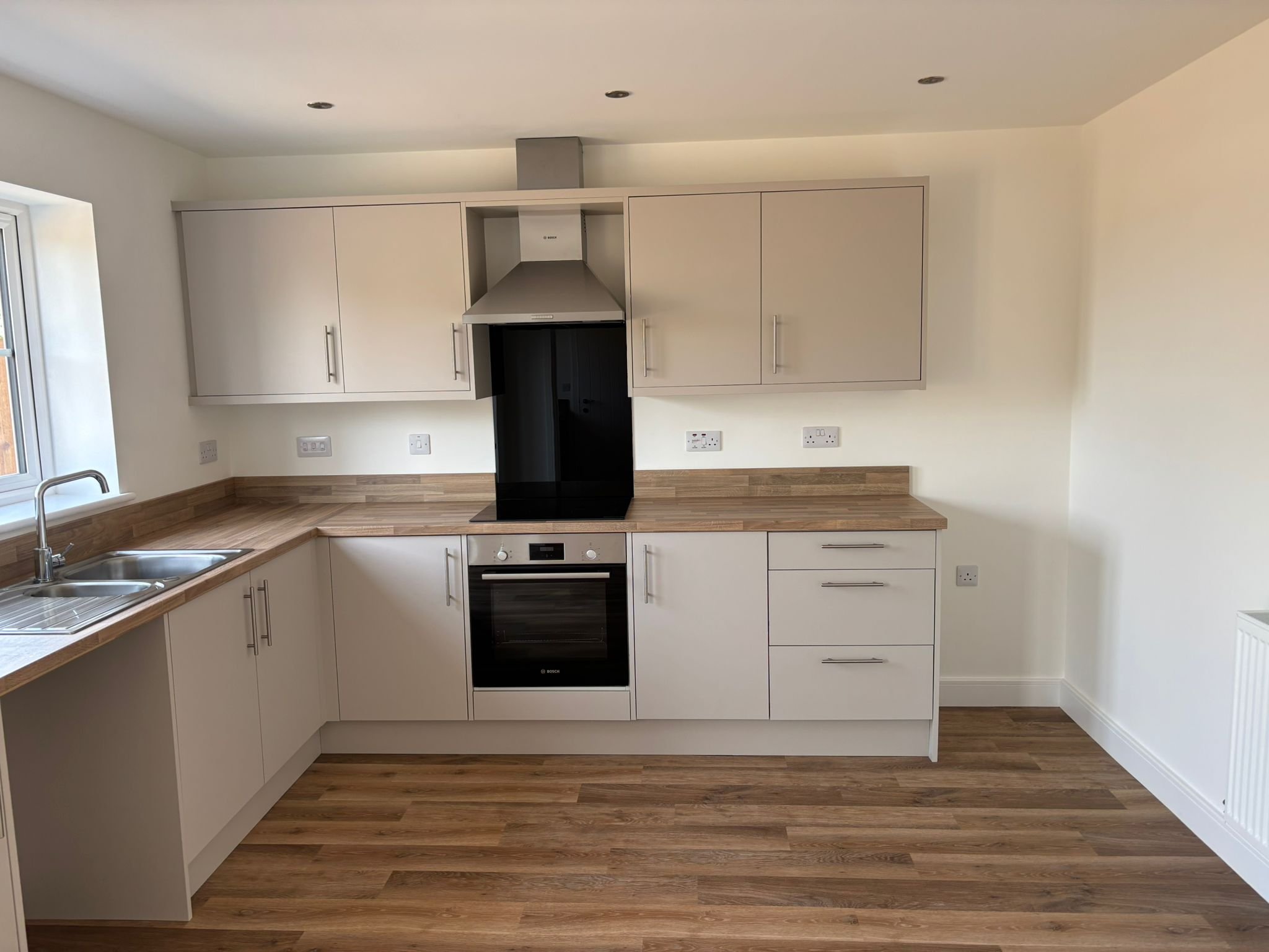 Modern kitchen with white cabinets, wooden countertops, and wood flooring. Features a built-in oven, stovetop, double sink, and electrical outlets on the wall.