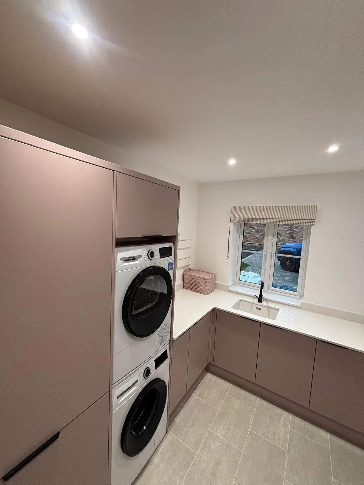 A modern laundry room with a stacked washer and dryer, beige cabinets, a small window with a striped valance, and a white countertop with a sink.