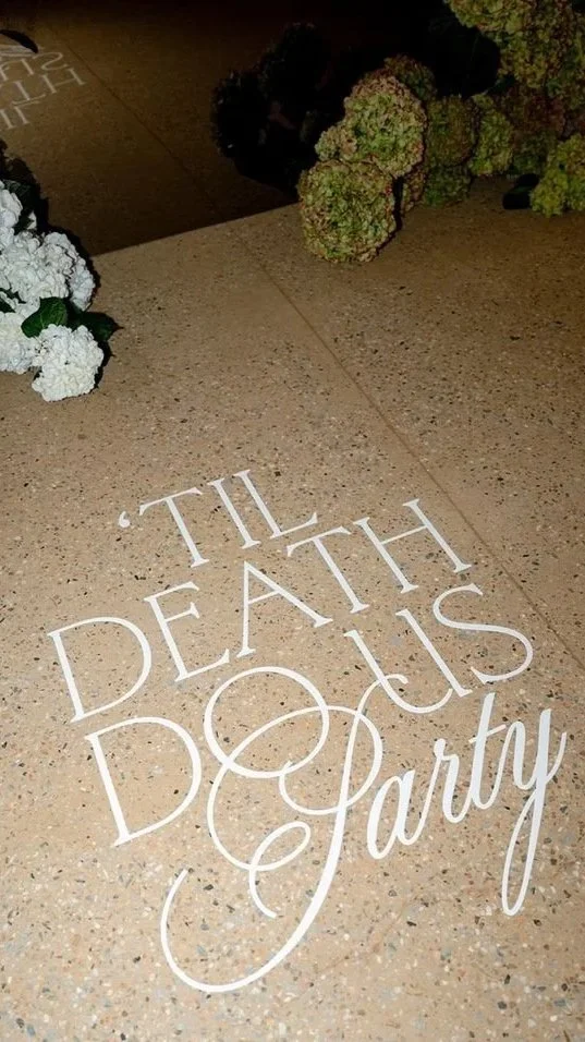 Decorative writing on the floor reading 'Till Death Do Us Part' with floral arrangements nearby.