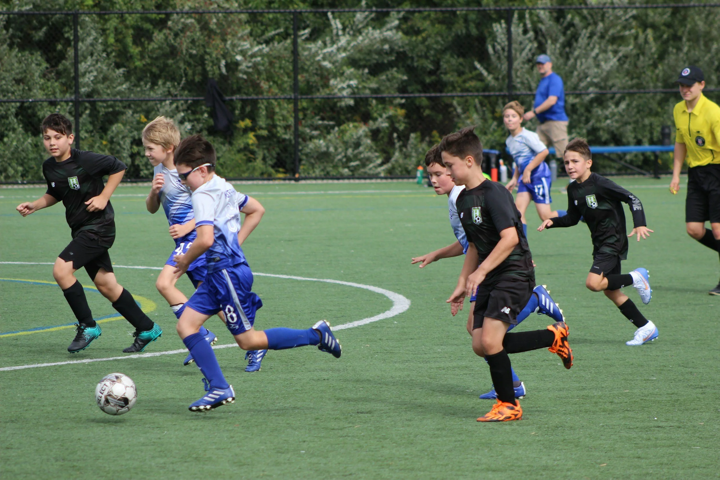 Young boys playing soccer on a green field during daytime, wearing black and blue uniforms with white and green logos.