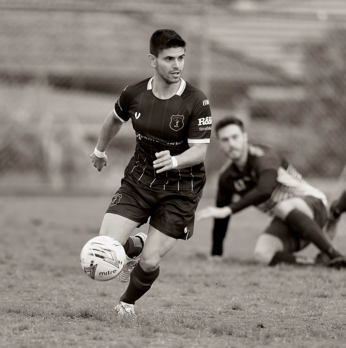 A soccer player in a black jersey and shorts running with a ball, while a goalkeeper is on the ground in the background during a game.