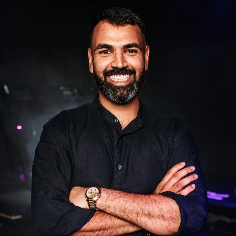 Man with dark hair and beard, smiling, wearing a black shirt with sleeves rolled up, crossed arms, dark background with blurred lights.