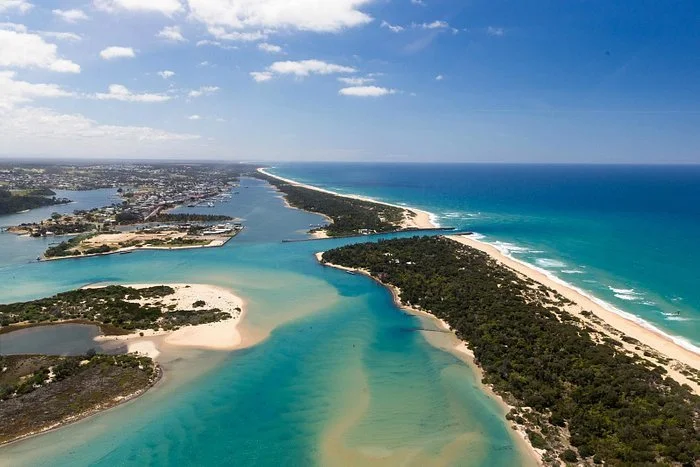 Aerial view of a coastal area with a river and island systems leading to the ocean, featuring sandy beaches, greenery, and a city in the distance.