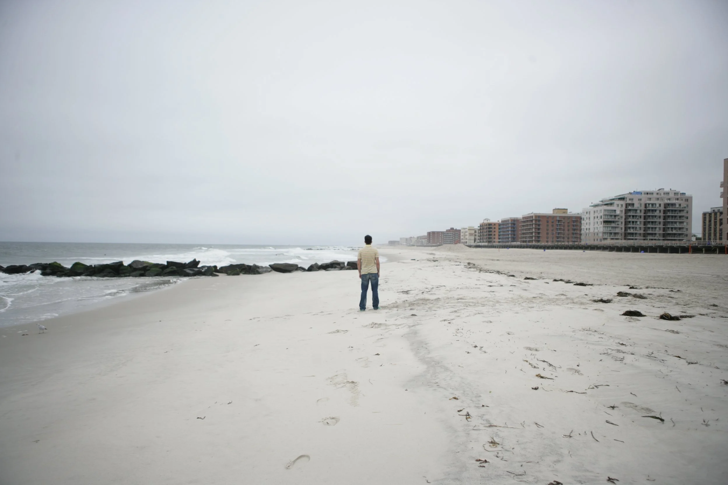 A solitary man in a striped shirt and jeans walking on a deserted beach with footprints in the sand, looking towards the ocean and a row of buildings on the horizon under an overcast sky.