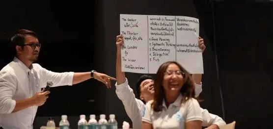 Three people at a presentation, one woman smiling and holding a large paper with writing, others pointing and looking at her, bottles on table.