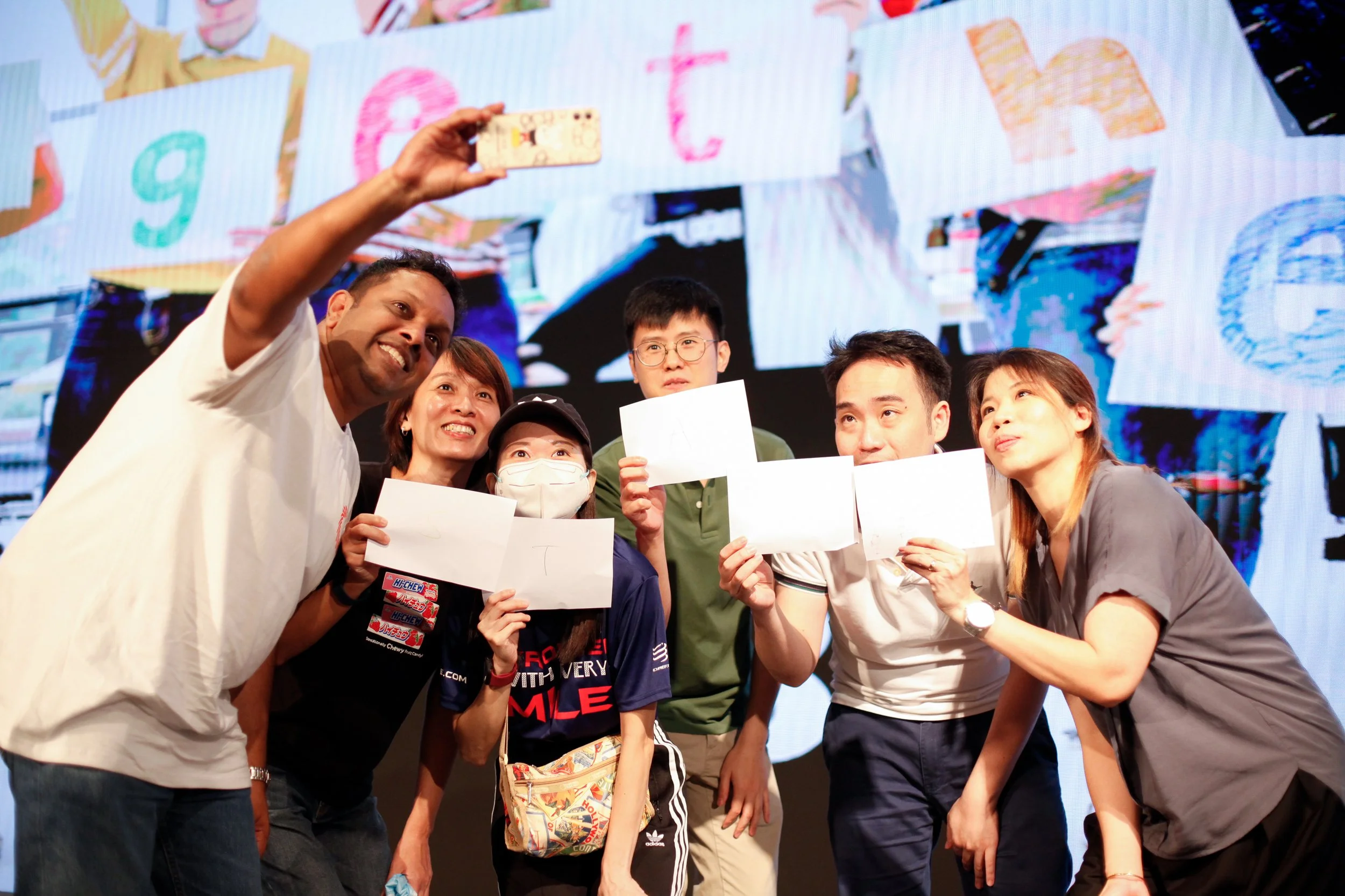A group of six diverse people taking a selfie together, with some holding blank papers, on a stage with colorful artwork in the background.