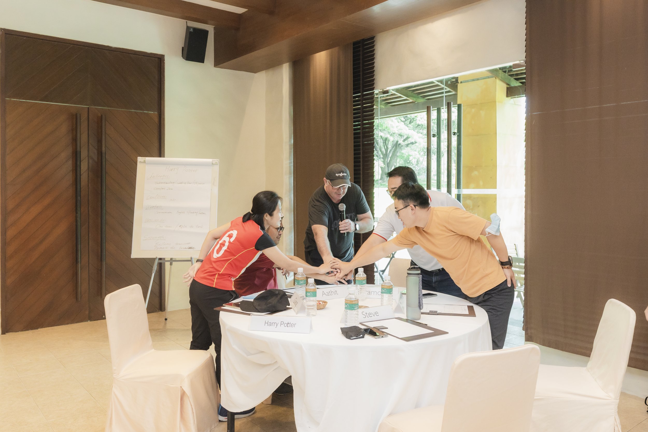 Group of five people in a meeting, with four stacking their hands and a man holding a microphone. They are around a round table with water bottles and name tags, in a room with glass doors and a display board.