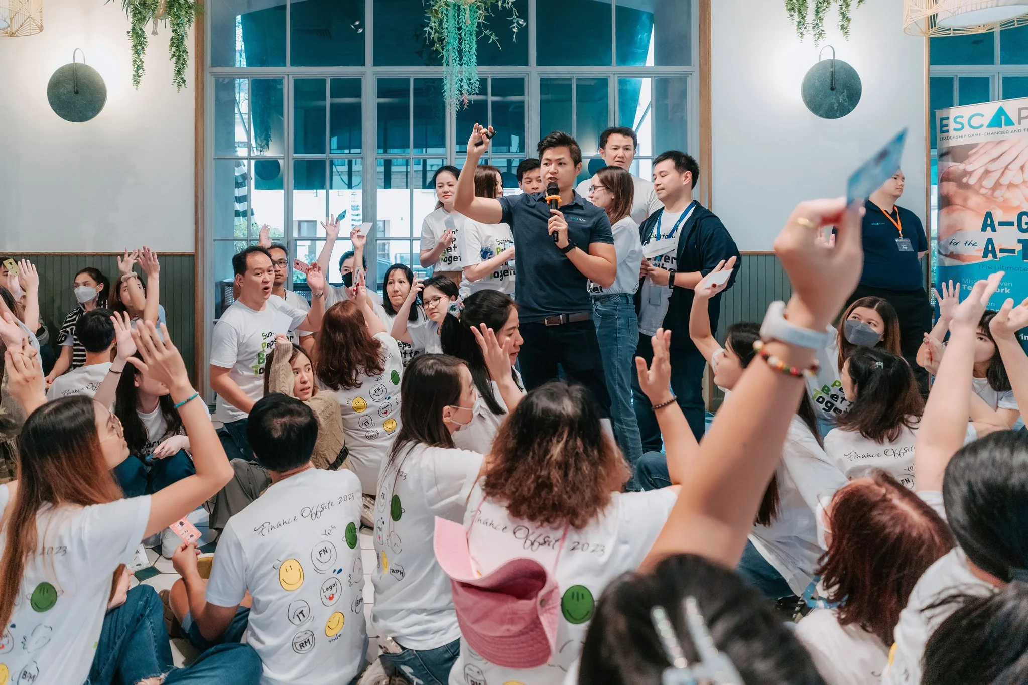Group of people gathered indoors, some wearing white t-shirts with emoji prints, raising hands, listening to a man standing on a chair with a microphone, who is speaking or giving a presentation.