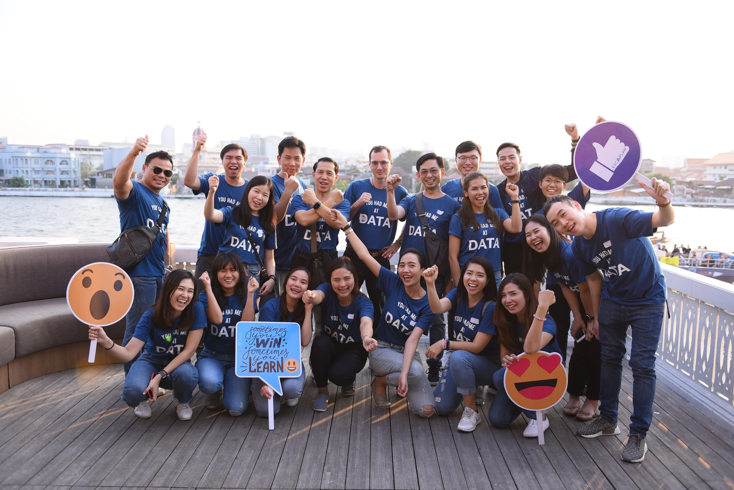 Group of people wearing matching blue shirts celebrating outdoors near a body of water, holding emoji and sign props, smiling, raising fists, and making victory gestures.