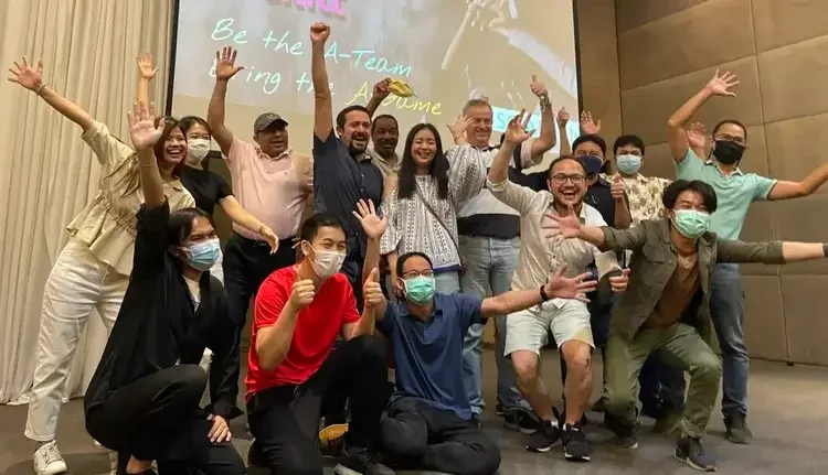 Group of diverse people celebrating on stage with raised hands, some wearing masks, in front of a screen displaying text.