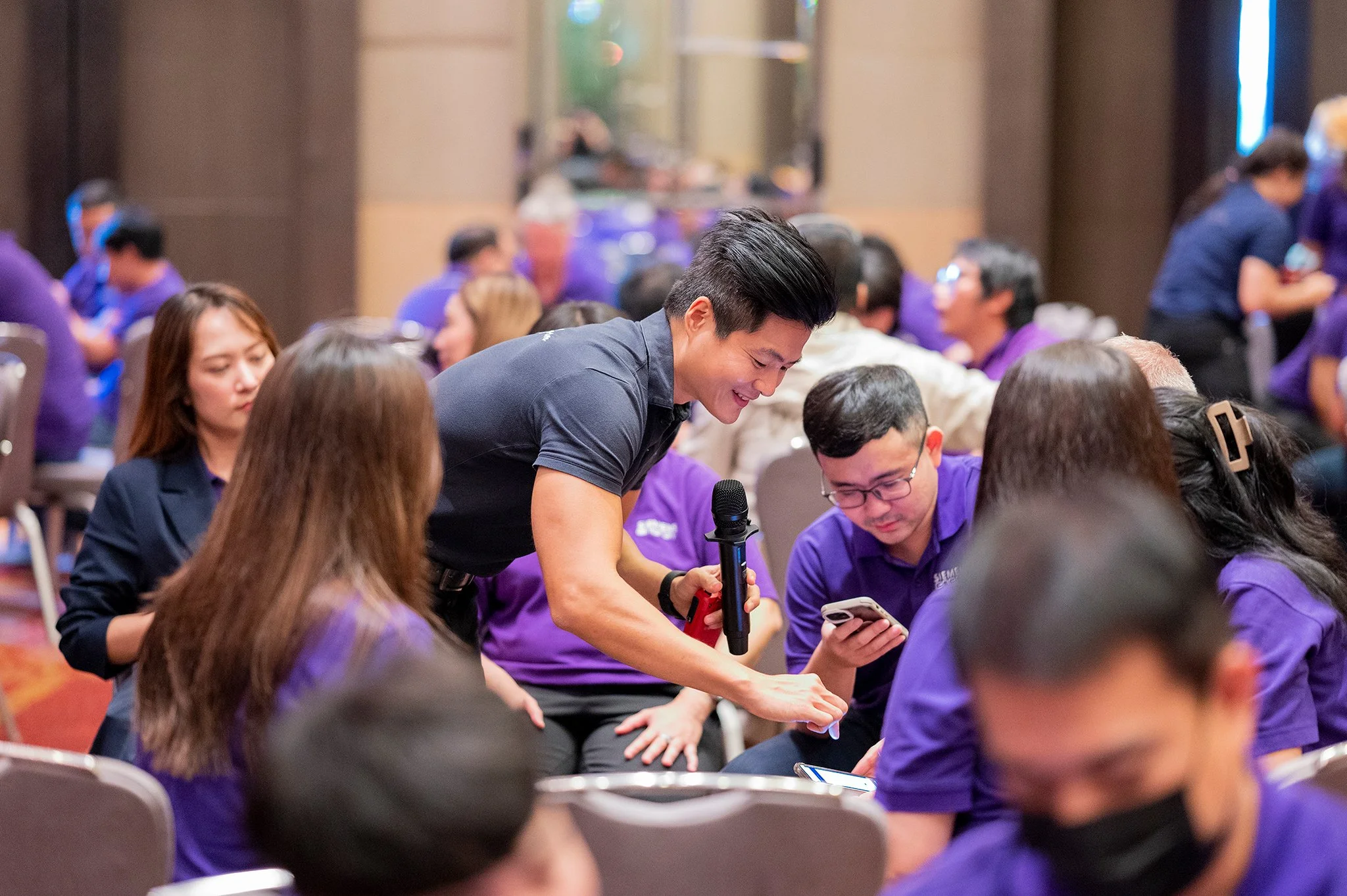 People sitting around tables at a conference or event, with one man standing and smiling while holding a microphone and showing something on his phone to the group.