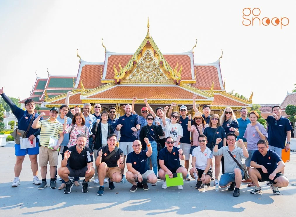 A group of people posing cheerfully in front of a traditional Thai temple, some raising their fists or waving, on a sunny day.