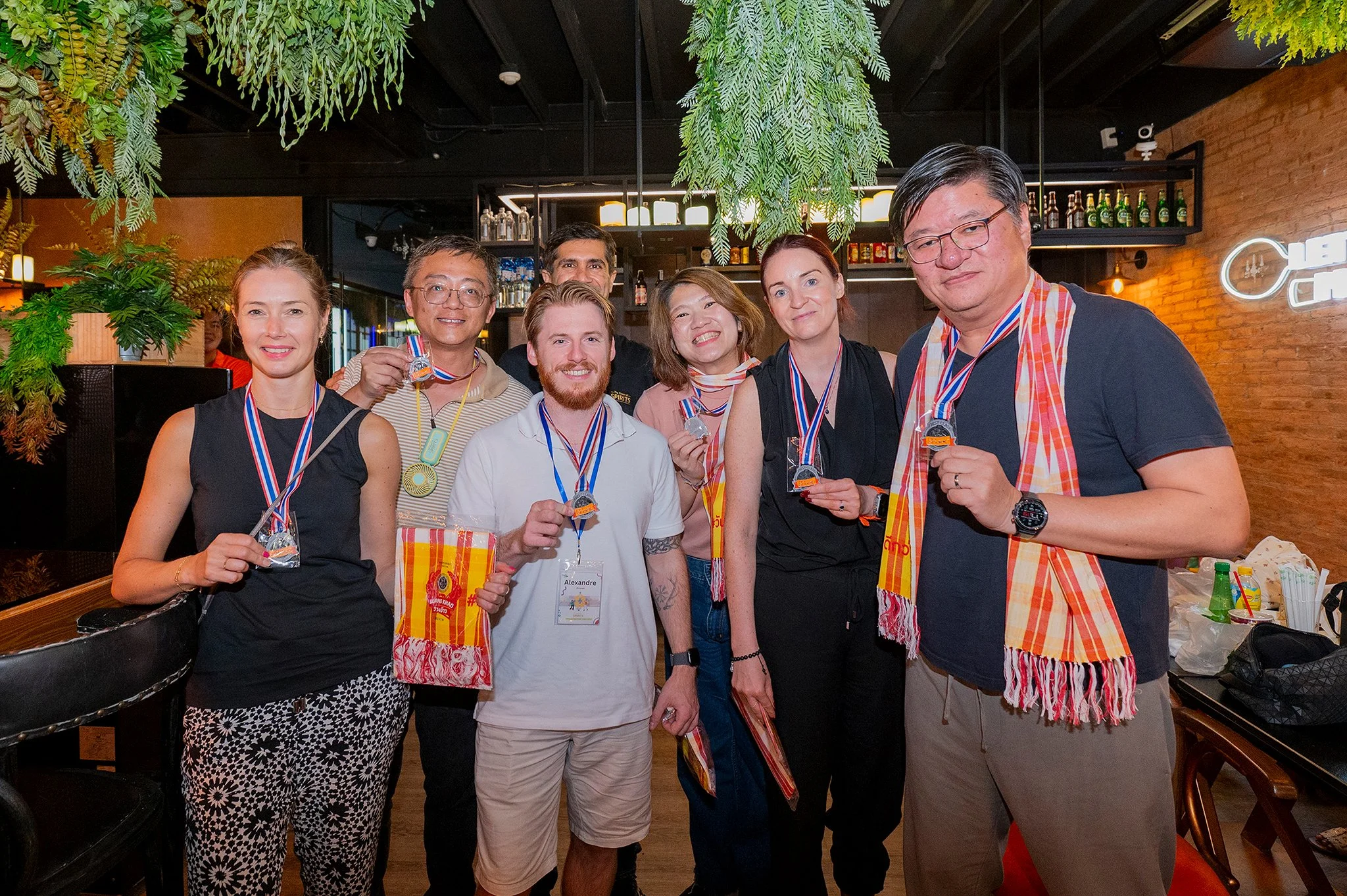 Group of people at an indoor celebration event, wearing medals and scarves, smiling, with a bar and decorative greenery in the background.