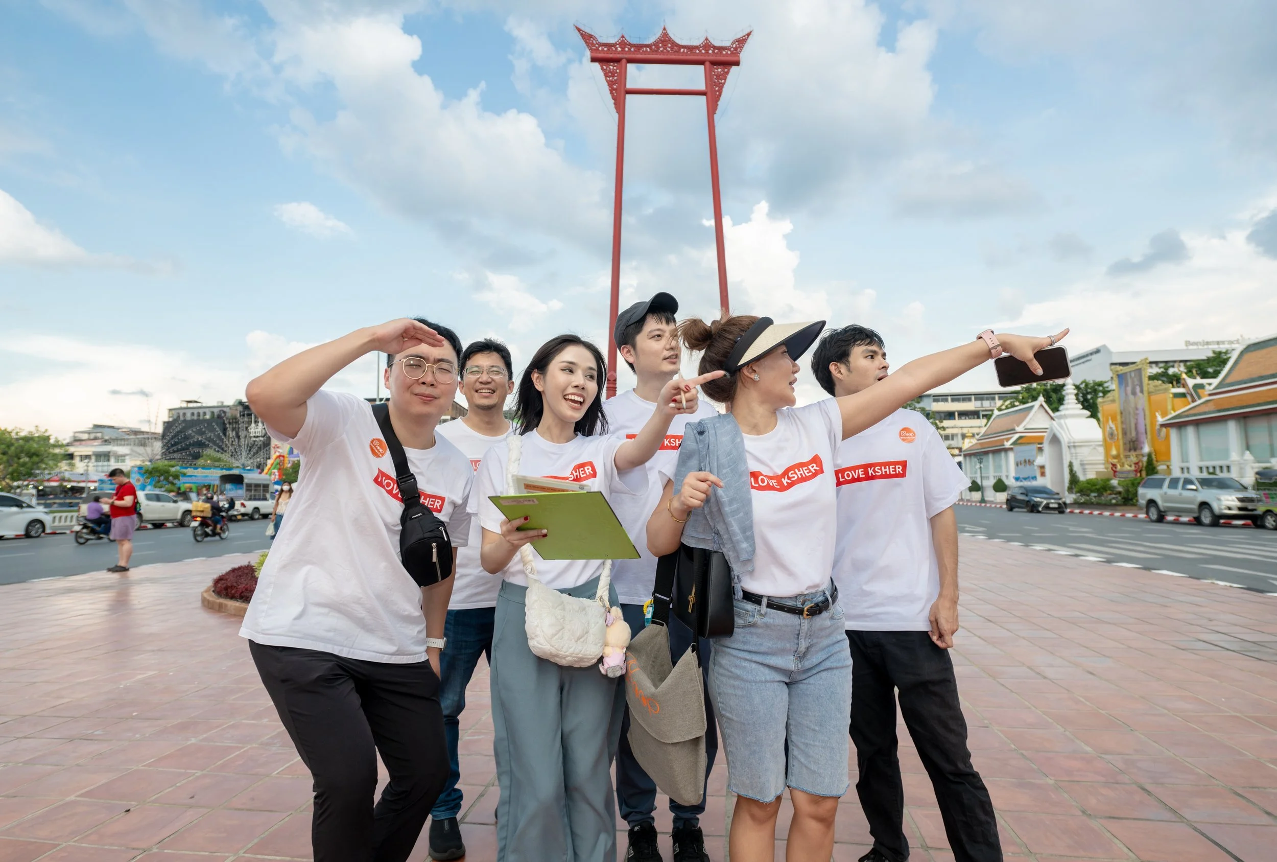 Group of six people taking a selfie in front of a large red monument in a city square, with traditional Thai buildings and cars in the background.