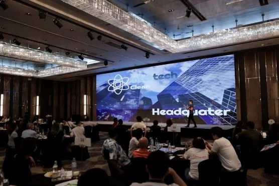 A conference with a large screen displaying a logo and the words "Headwater." The room is filled with seated attendees, and the venue has modern lighting and decor.