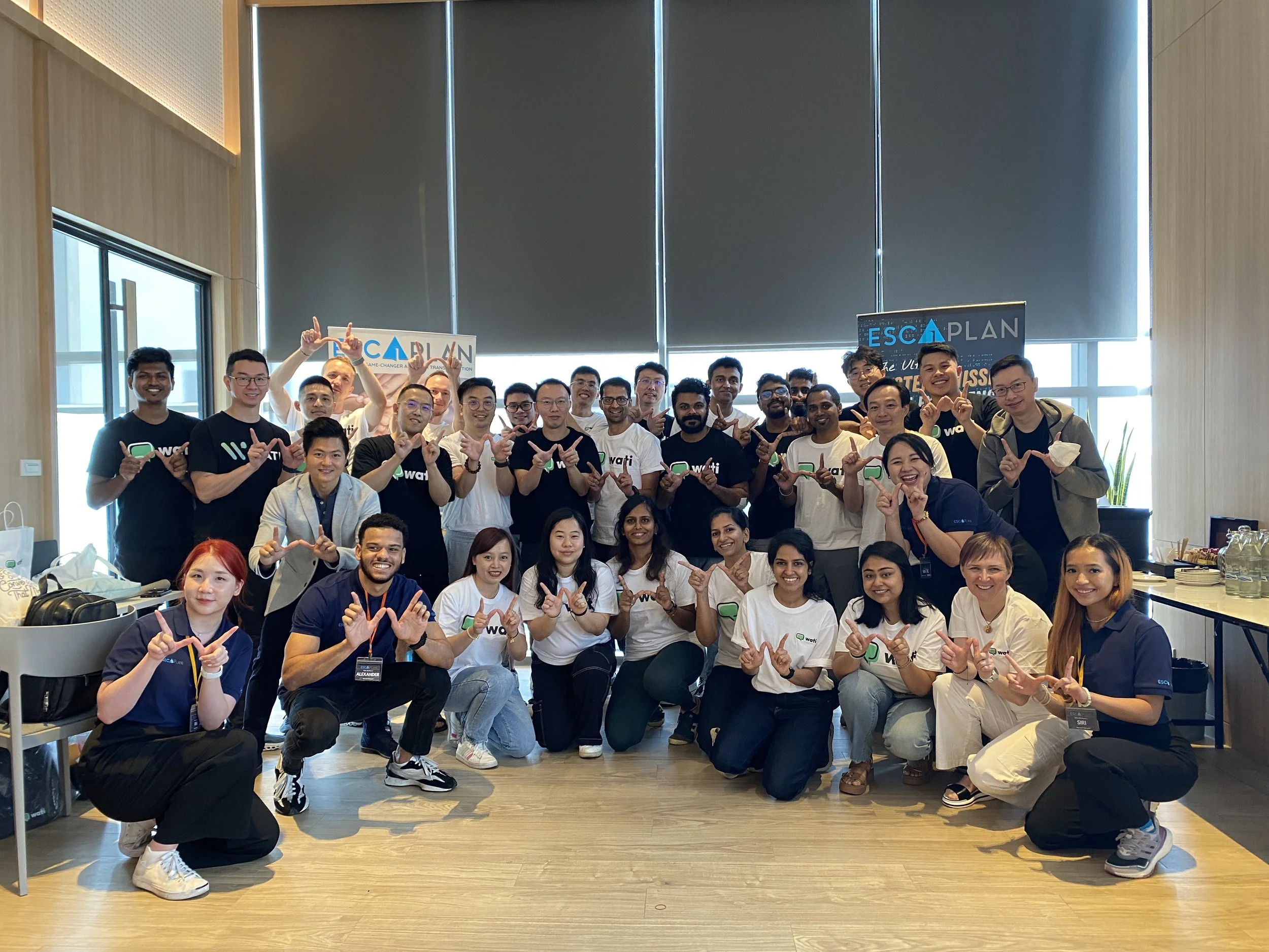 Group of people posing for a photo in an indoor space, some holding signs that say 'ESCAPLAN'. They are smiling and making hand gestures.