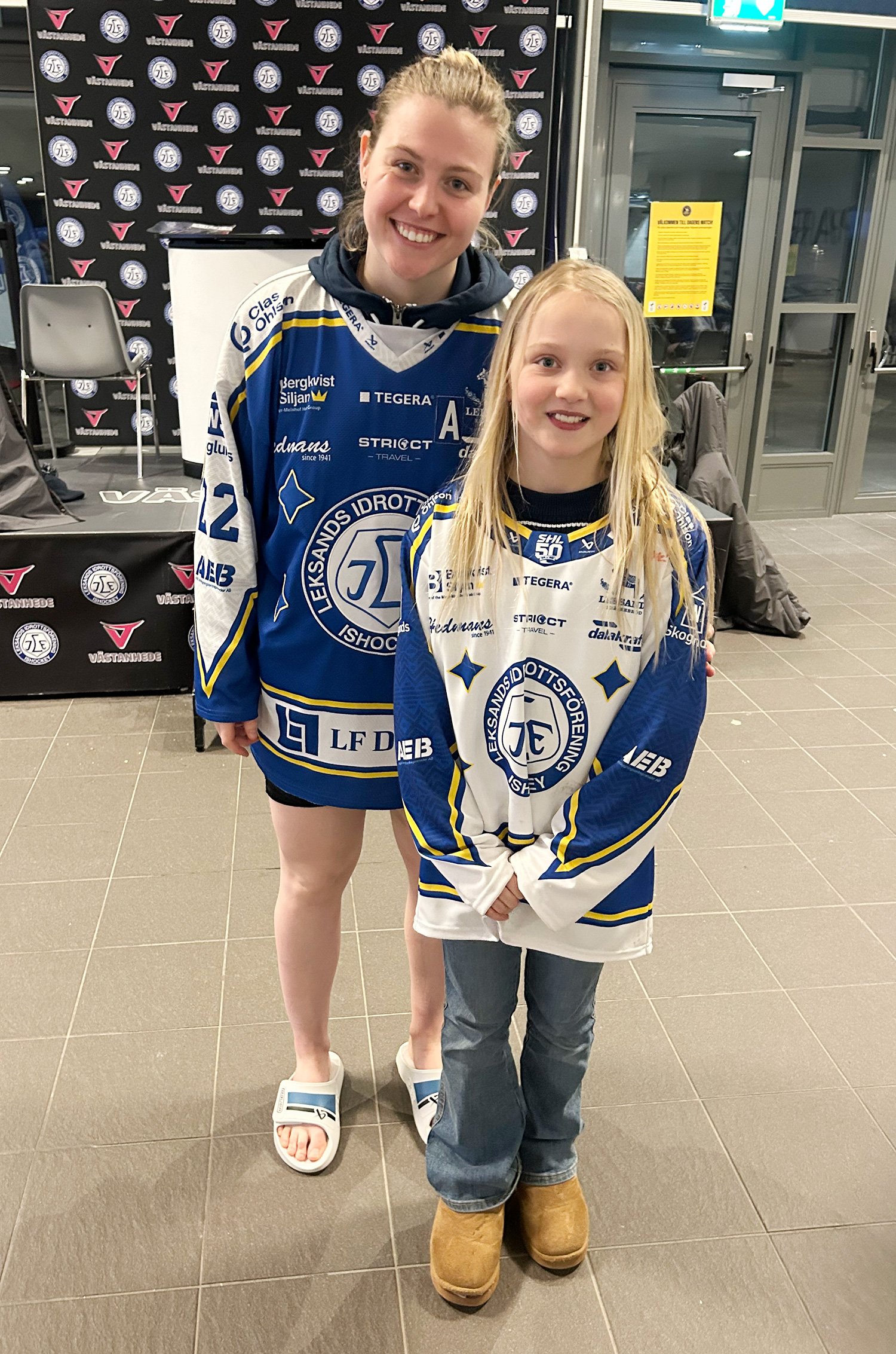 Två unga tjejer klädda i blå och vit sporttröjor från Leksands IF hockeyklubb, står inne i en byggnad, en av tjejerna är barfota och den andra bär Uggs, de ler mot kameran.