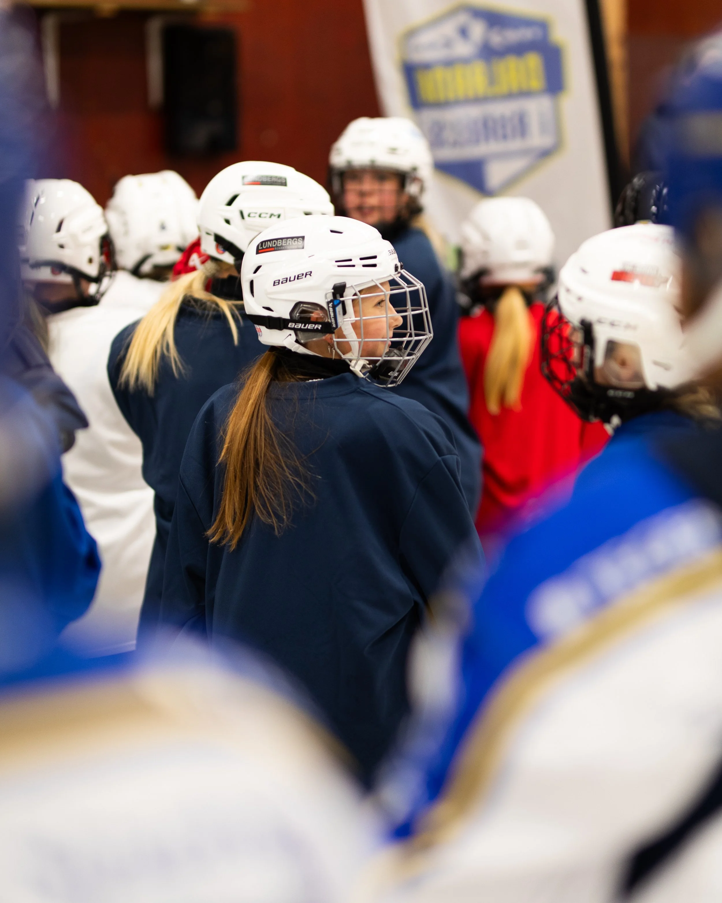 Ung hockeyspelare i vit hjälm och blåa kläder, förmodligen på isrinken, med en grupp andra hockeyspelare i bakgrunden.