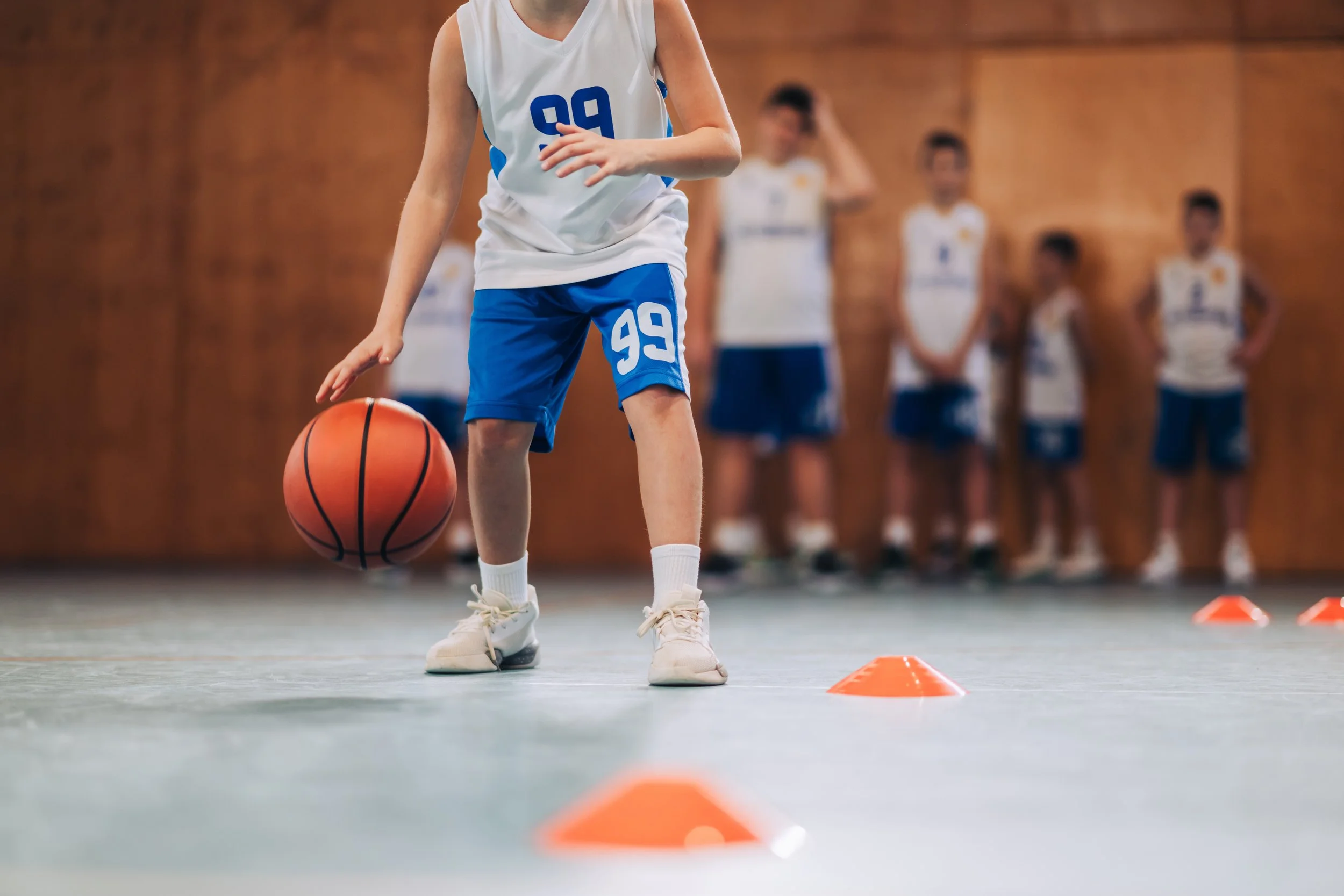 Ung basketspelare dribblar en basketboll mellan orange koner på en inomhusbasketplan, med tre andra barn i bakgrunden.