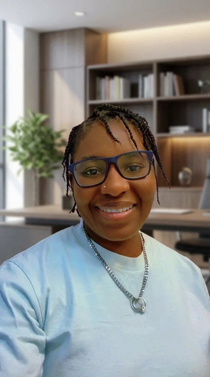 A smiling woman wearing glasses and a white T-shirt, sitting in a modern office with a bookshelf and a plant in the background.