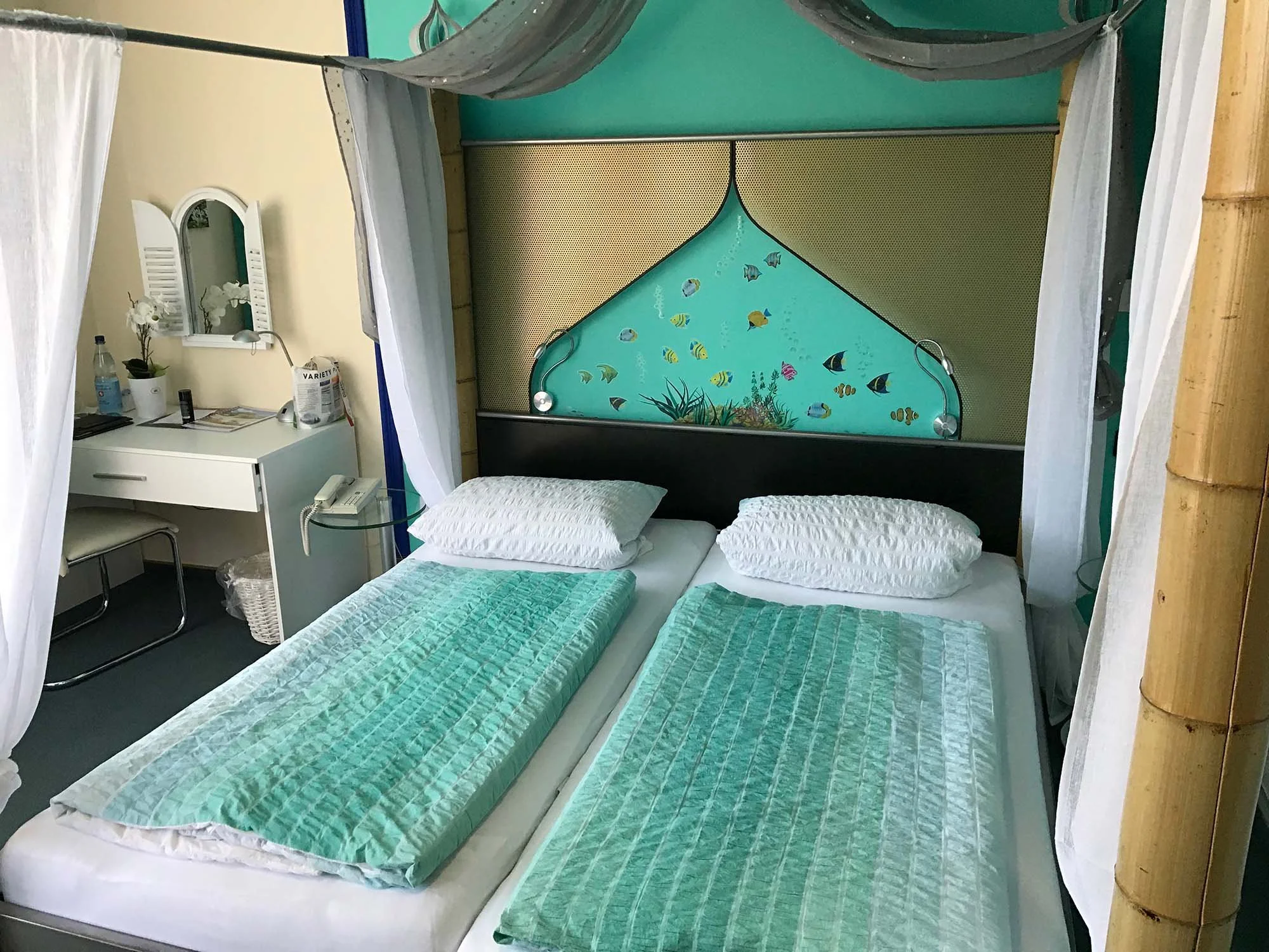 Ein Hotelzimmer mit vier auseinanderstehenden Betten mit grünen Bettdecken, einem Nachtschränkchen, einem Spiegel über einem Schreibtisch, einer Wasserflasche, einem Telefon und einem Stuhl. Die Wand hinter den Betten ist mit einem Wandbild von einem